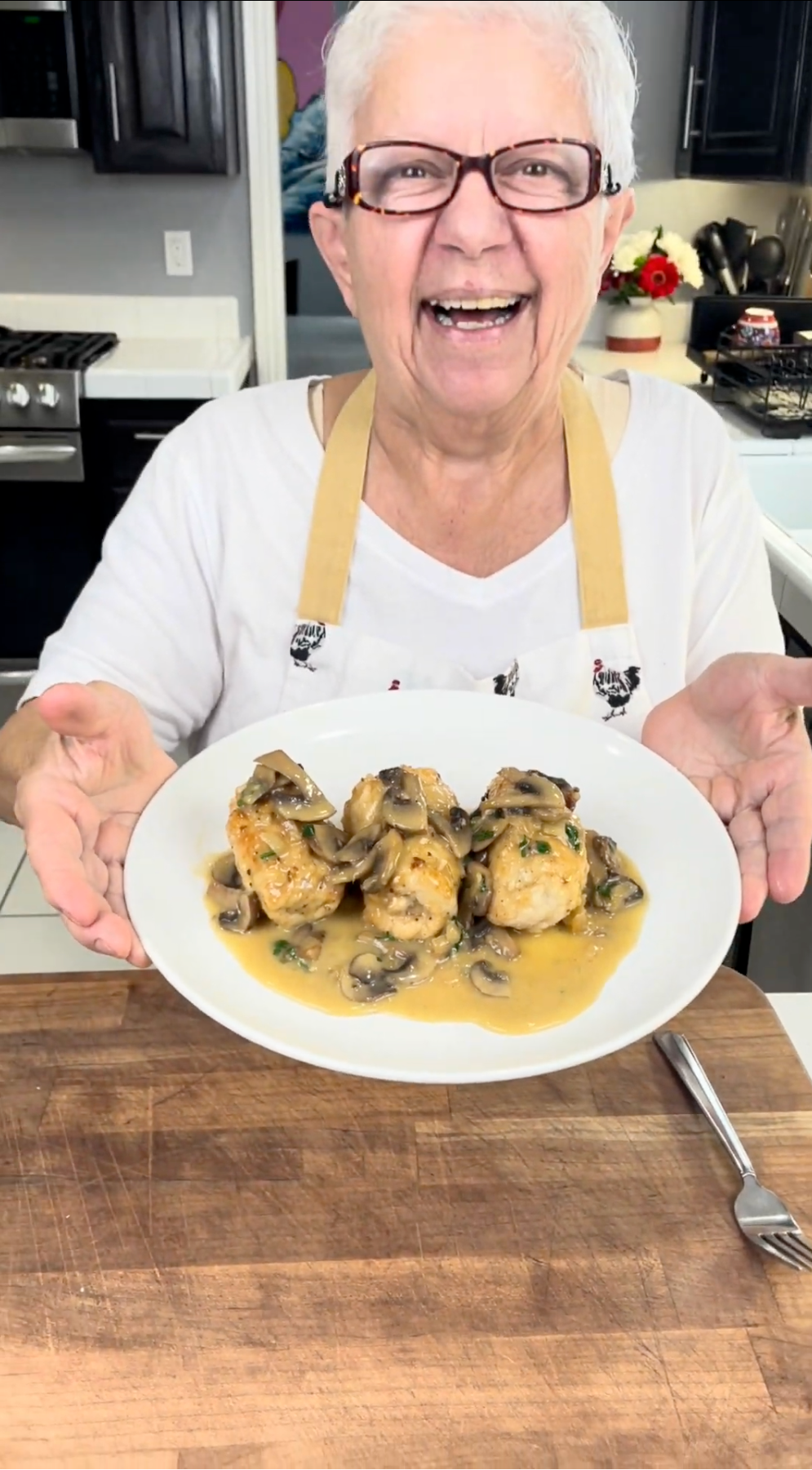 An elderly woman with white hair, glasses, and an apron smiles while holding a plate of chicken with mushroom sauce in a bright kitchen.