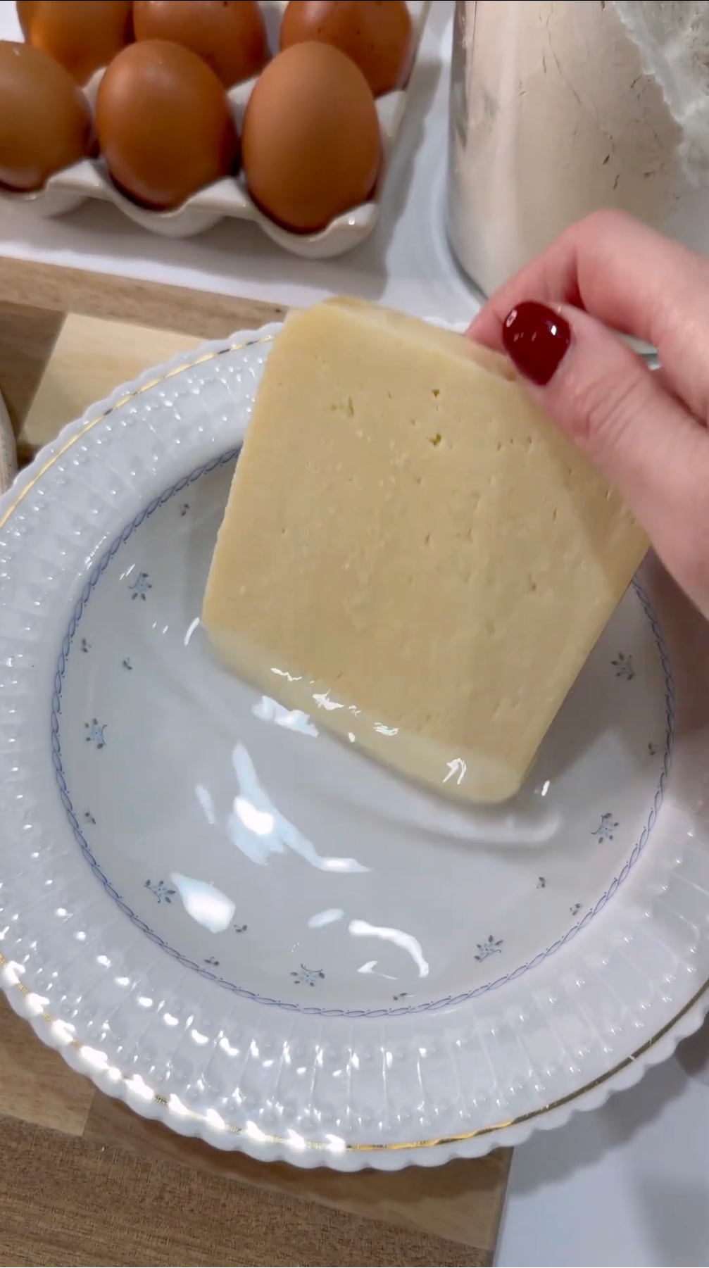 A hand with red nail polish holds a wedge of cheese over a patterned white plate; eggs and a flour container are visible in the background.