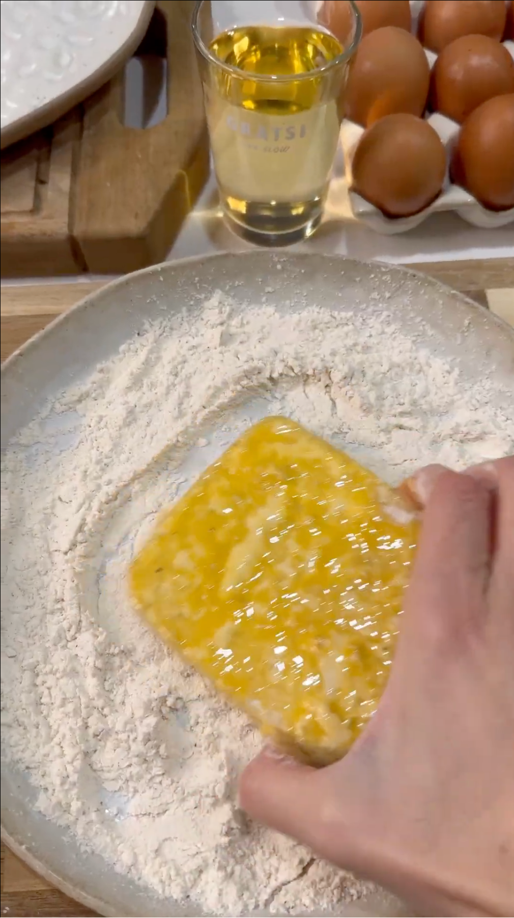 A hand coats a slice of bread in beaten egg over a plate of flour. A tray of eggs, a glass of oil, and a wooden cutting board are visible in the background.