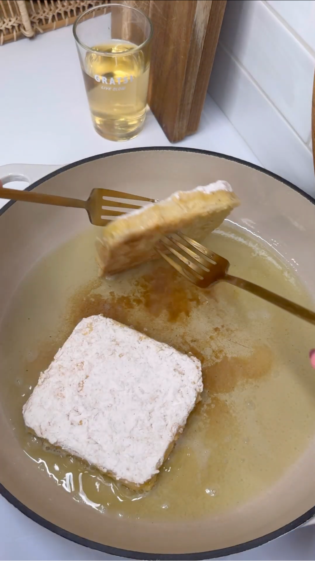 A square piece of breaded food is being lifted with a gold spatula from a pan with oil, while another piece fries in the oil. A glass of yellow liquid sits on the counter in the background.
