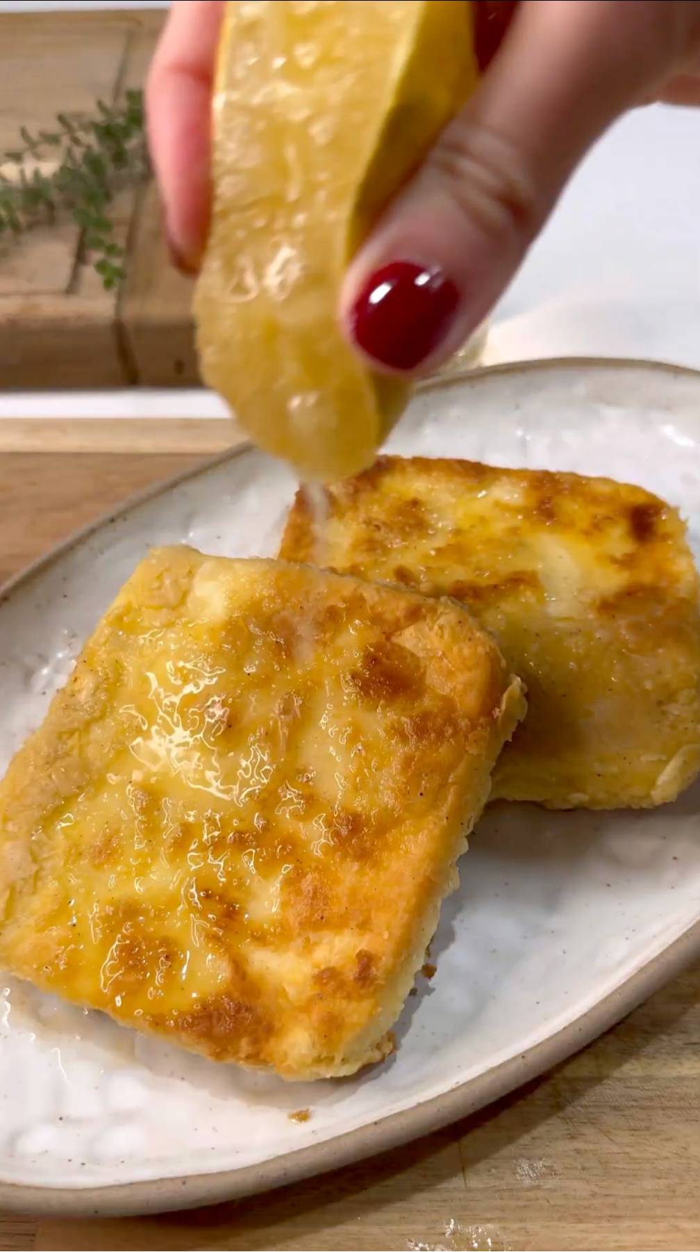 Two golden, crispy slices of fried food on a white plate, with a hand squeezing a lemon wedge over them, drizzling fresh juice on top. The scene suggests a fresh, homemade meal.