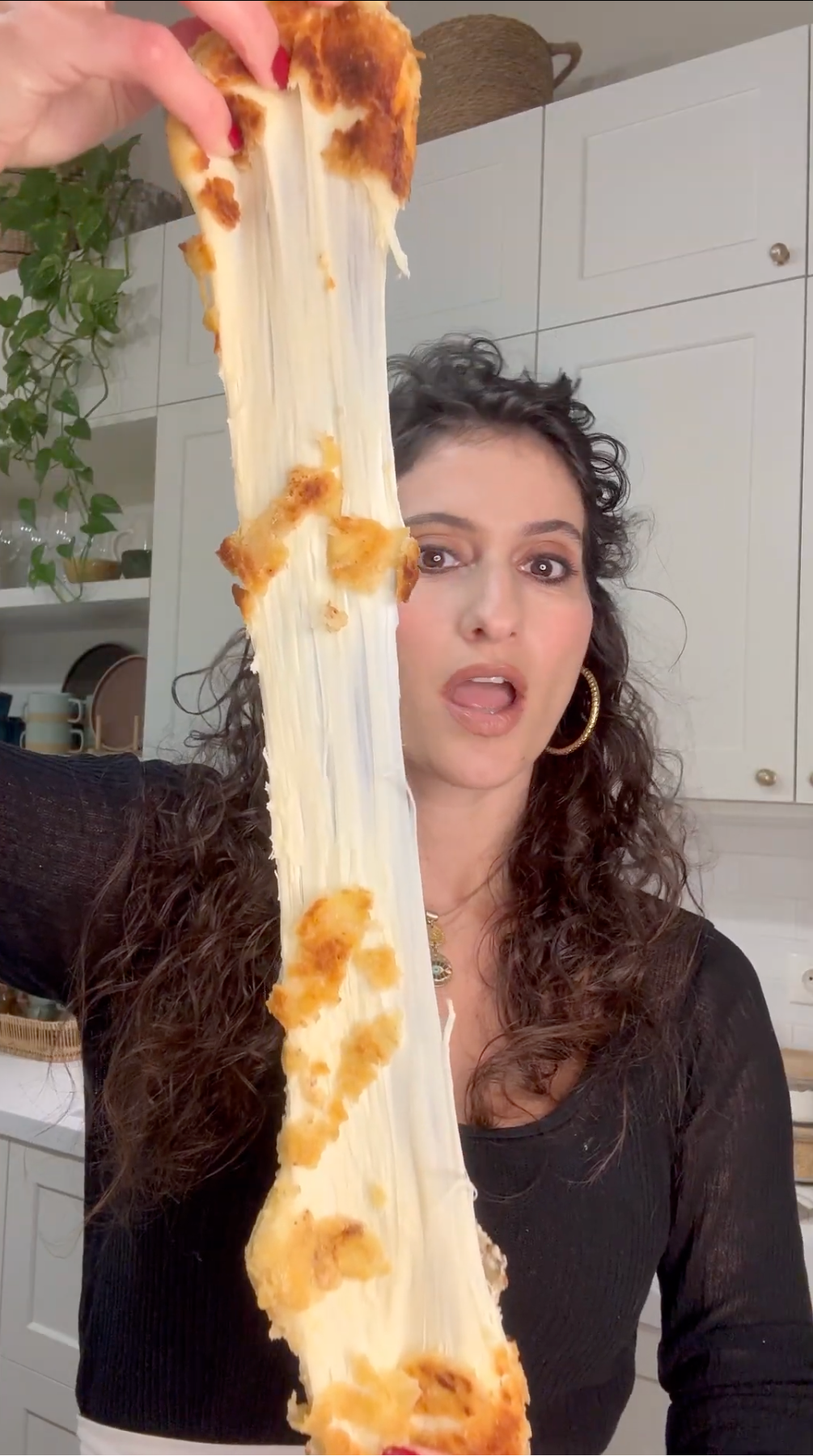 A woman in a kitchen holds up a large, stretchy piece of melted cheese with bits of breading, stretching it vertically above her head. She looks surprised or impressed by the length of the cheese pull.