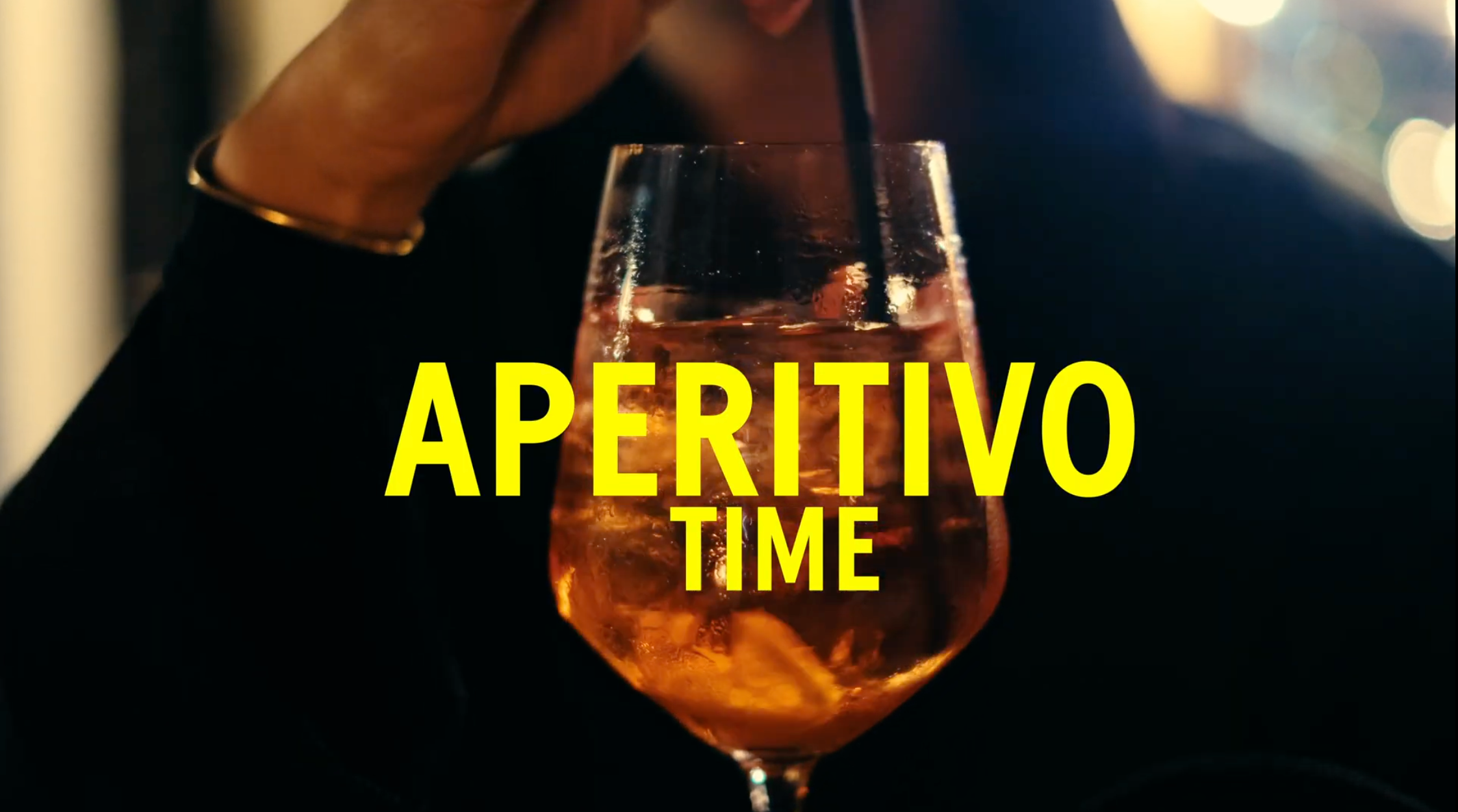 A large glass filled with an orange beverage and ice sits in front of a person drinking with a straw. Bold yellow text over the glass reads Aperitivo Time.