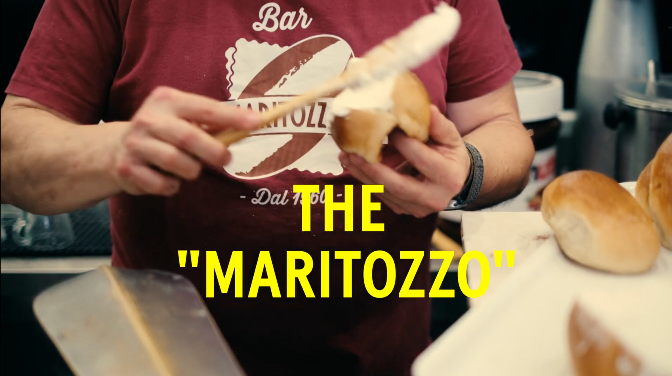 A person spreads cream inside a sliced bun while wearing a maroon shirt with Bar Maritozzo on it. Text in yellow reads: THE MARITOZZO.