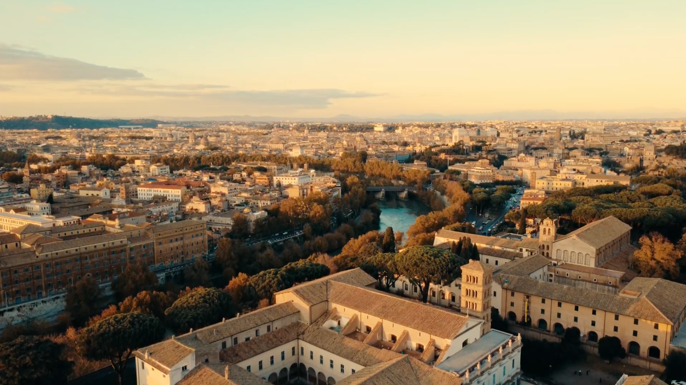 Aerial view of a city with historic buildings, terracotta rooftops, and tree-lined streets at sunset, with a river running through the center and distant hills under a partly cloudy sky.