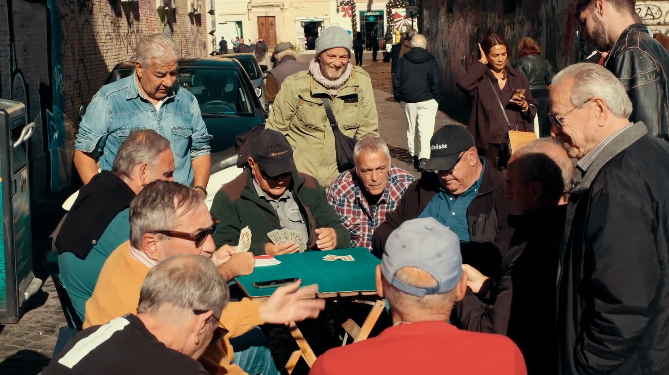 A group of older men sit around a table playing cards outdoors on a sunny day, while others stand and watch. The scene is set on a street with people and cars in the background.