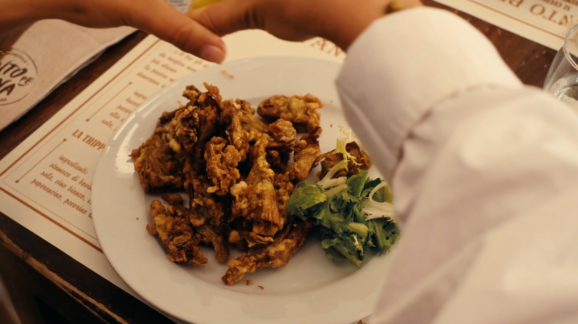 A person squeezes lemon over a plate of fried food with a small side of salad, set on a table with a menu underneath.