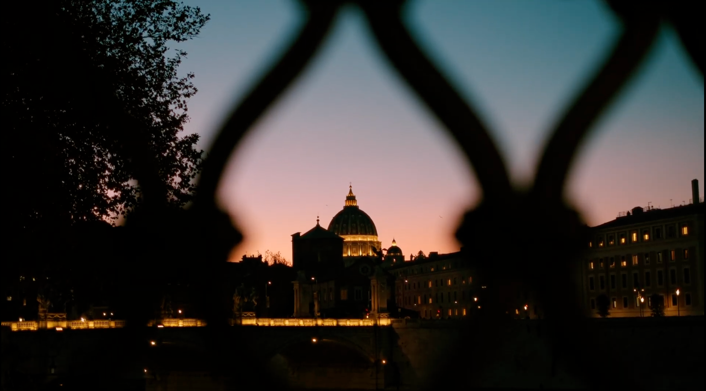 St. Peter’s Basilica at sunset, viewed through an ornate metal fence, with warm city lights and silhouettes of surrounding buildings against a colorful sky.
