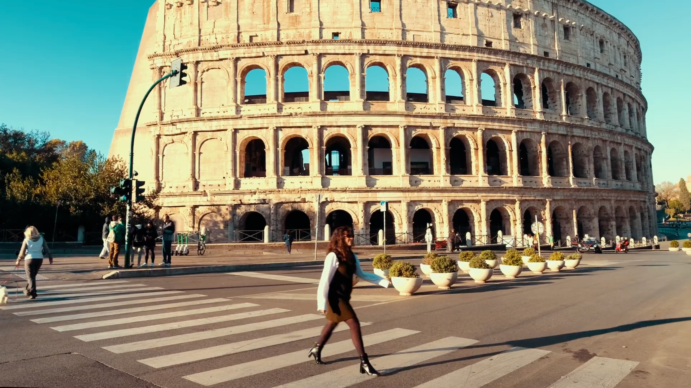 A woman crosses a street at a crosswalk in front of the Colosseum in Rome on a sunny day, while a few people stand nearby and cars are absent.