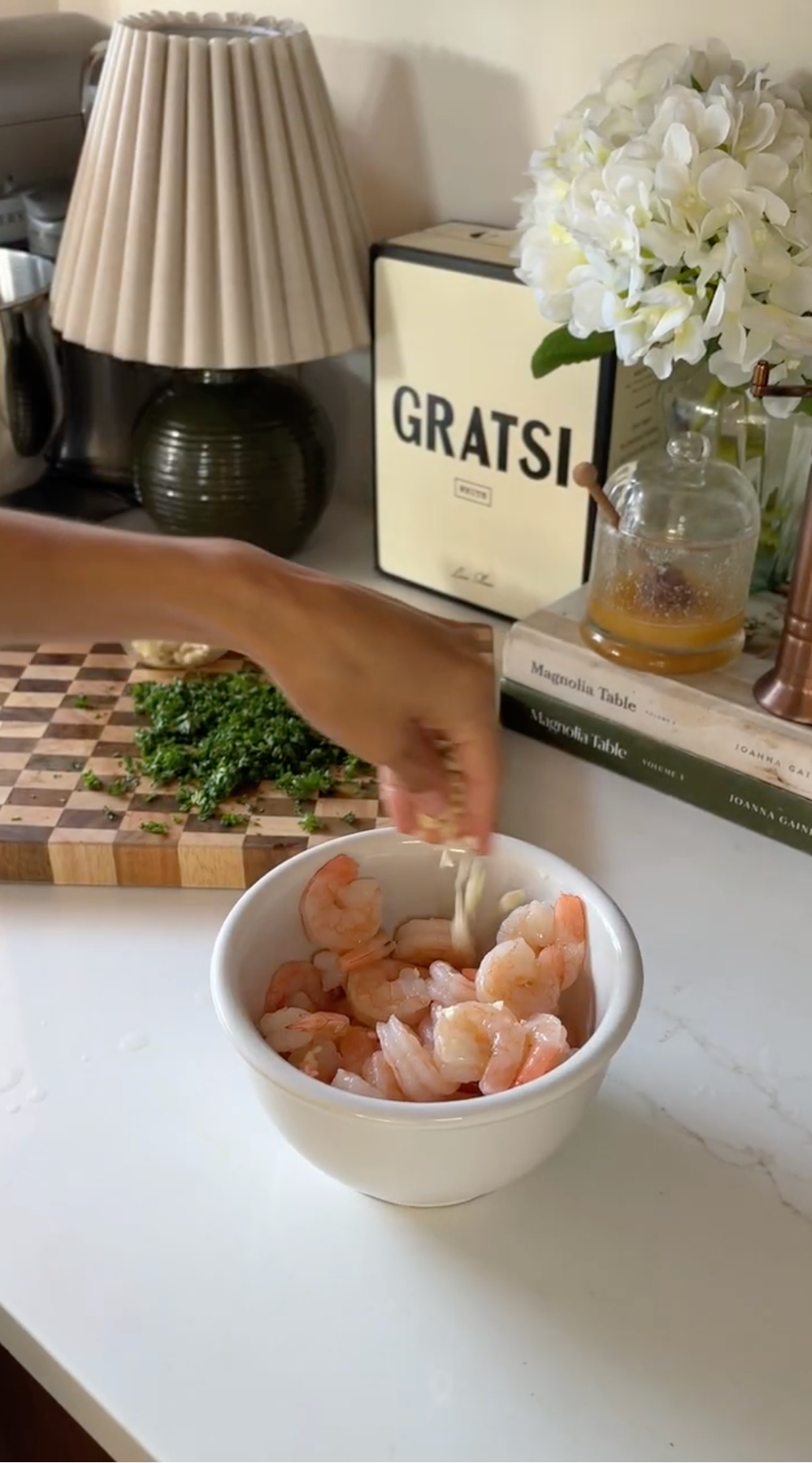 A hand adds chopped garlic to a white bowl filled with shrimp on a kitchen counter. In the background, there is a cutting board with chopped herbs, a lamp, books, flowers, and a box labeled GRATSI.