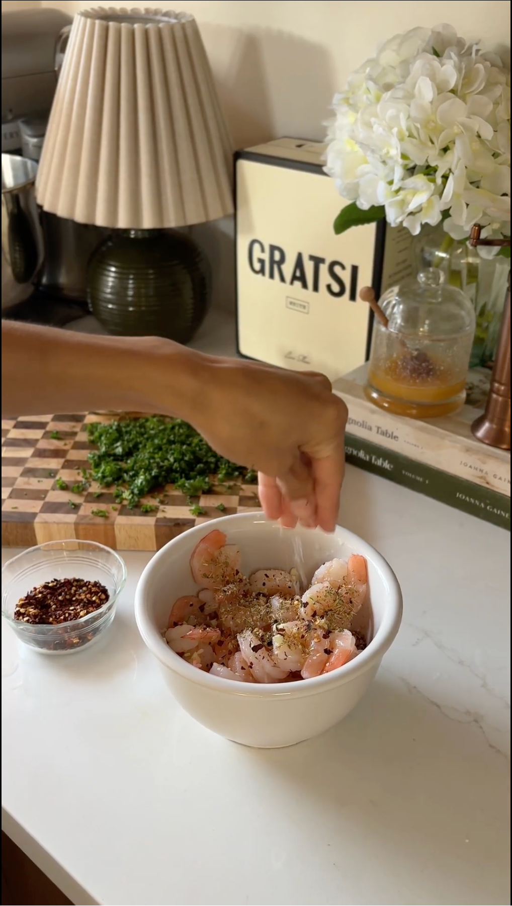A hand sprinkles seasoning onto raw shrimp in a white bowl on a kitchen counter, with a small bowl of red pepper flakes, chopped herbs on a cutting board, and a vase of flowers in the background.