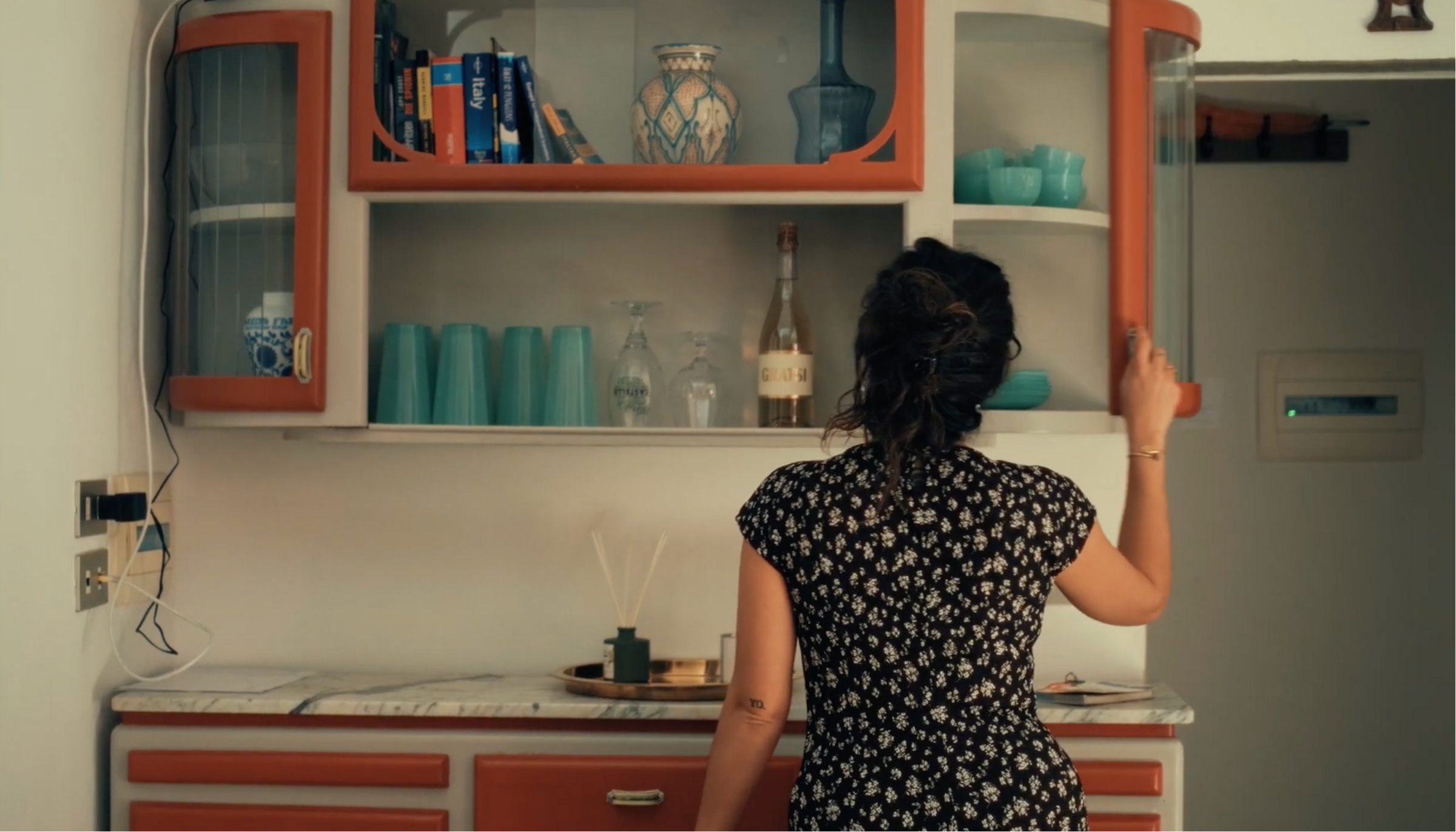 A woman in a black floral dress stands with her back to the camera, reaching to open a glass-fronted kitchen cabinet filled with blue dishes, glasses, a vase, and books above a countertop.
