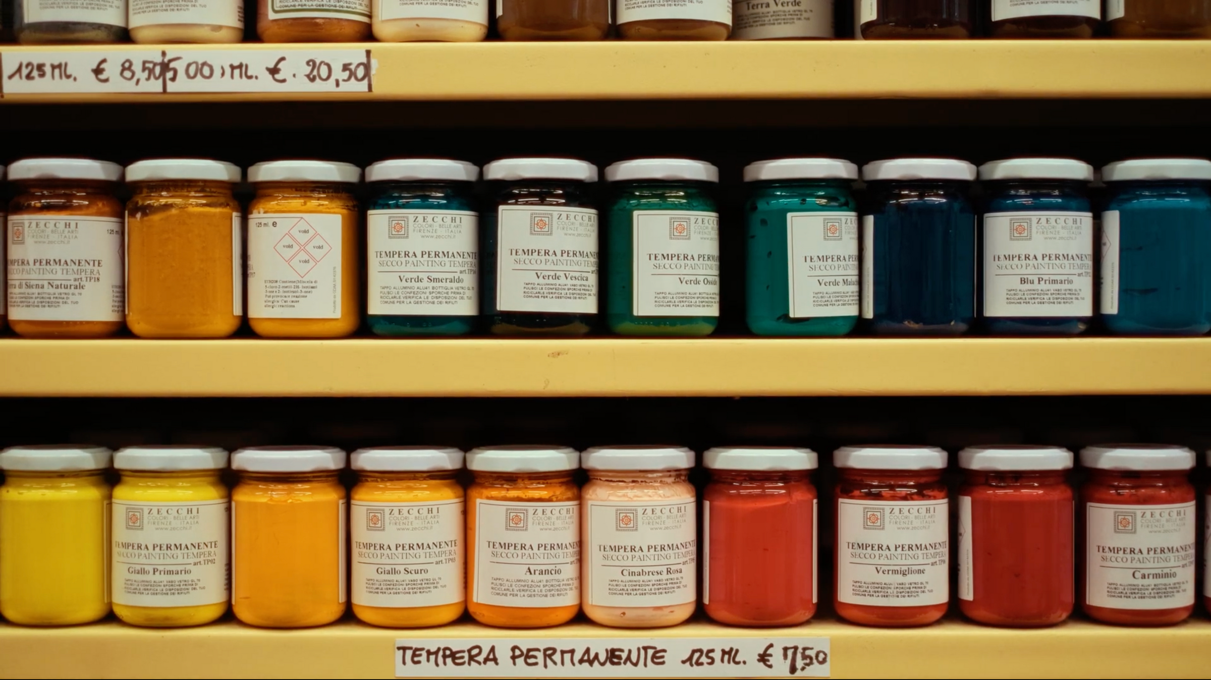 Rows of glass jars filled with colorful tempera paints are neatly arranged on shelves. Labels display paint names and colors like yellow, green, blue, and red. Price tags are visible above and below the shelves.