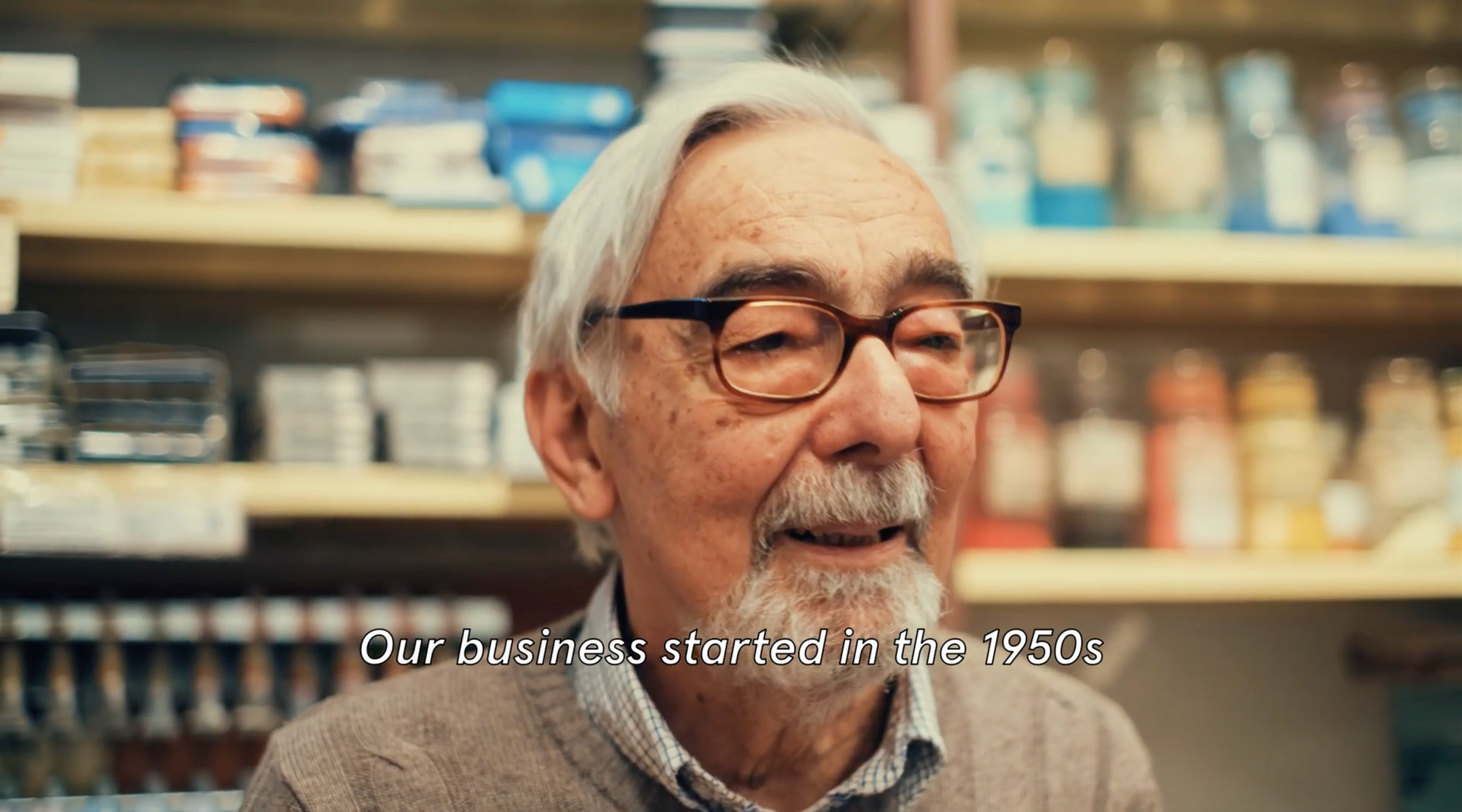 An elderly man with white hair, glasses, and a mustache smiles in a shop with shelves of products behind him. Text on the image reads, “Our business started in the 1950s.”.