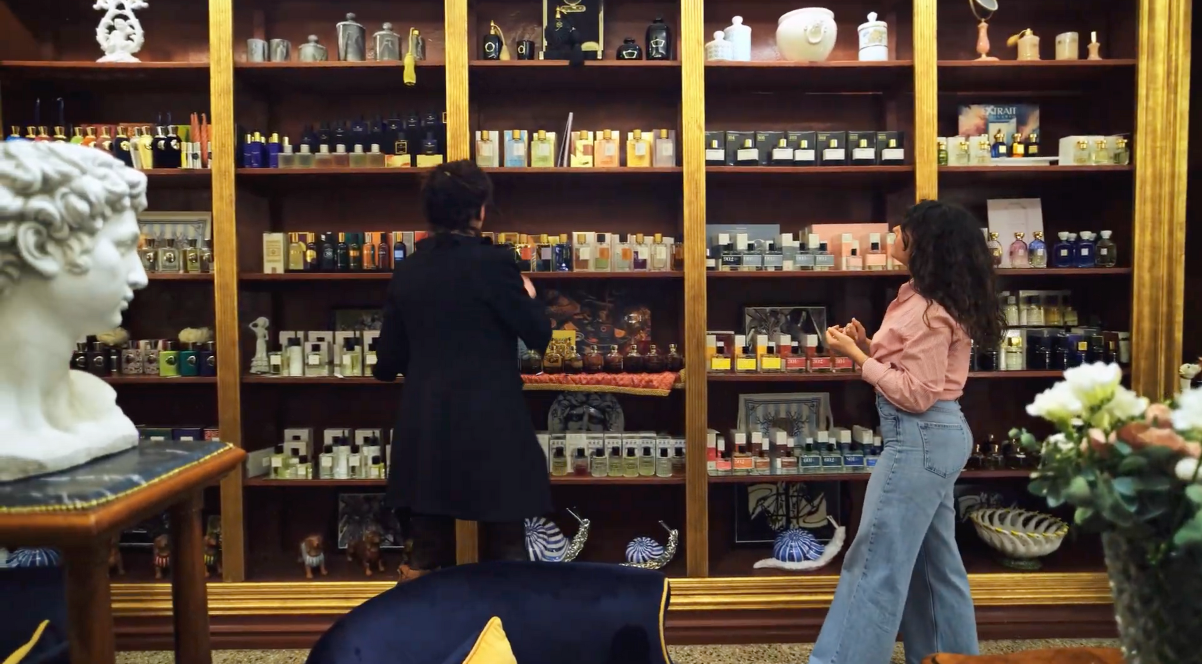 Two women browse a large, wooden shelving unit filled with various perfume bottles and boxes in an elegant boutique. A white statue and a vase of flowers decorate the foreground.