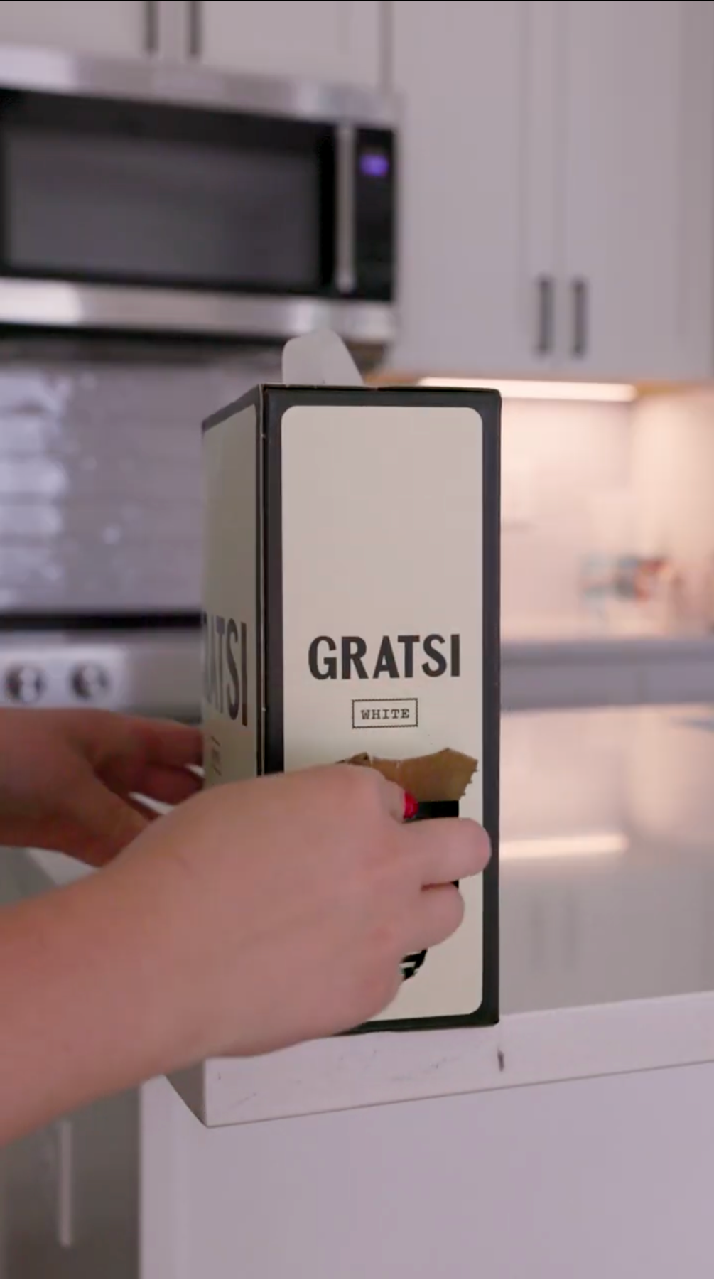 A person’s hands hold a boxed wine labeled “GRATSI” on a white kitchen counter, with white cabinets and a microwave in the background.