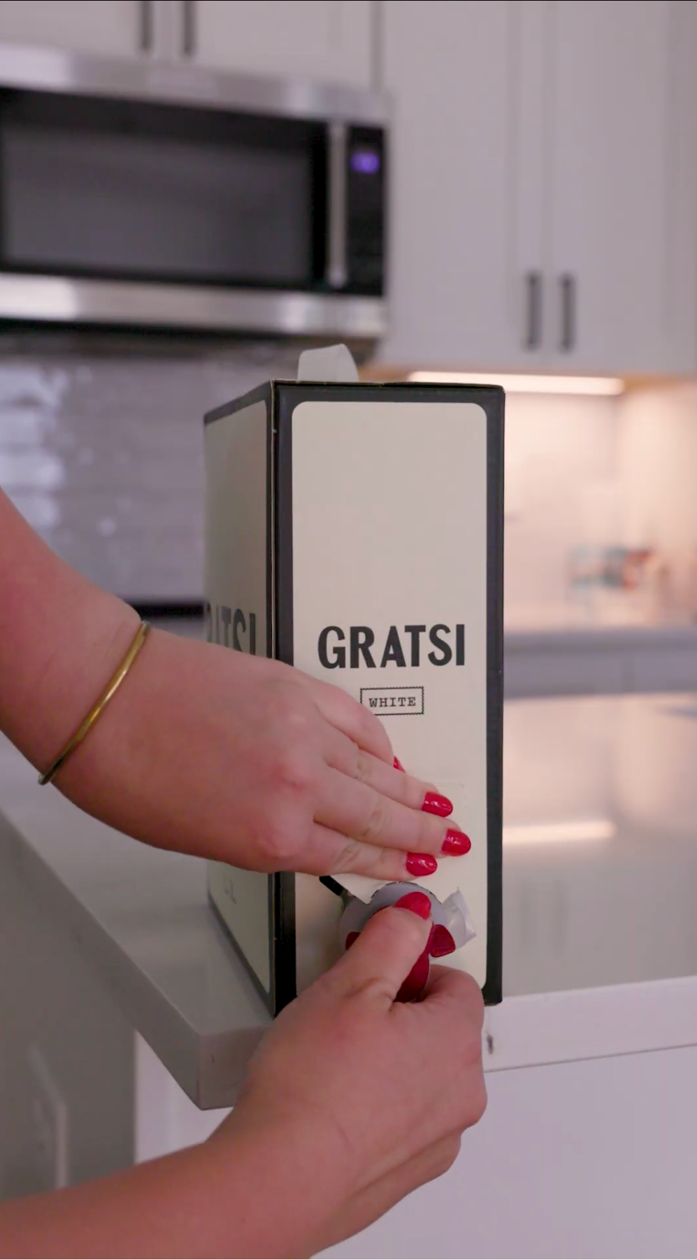 A person with red nail polish and a gold bracelet opens a box labeled GRATSI WHITE on a white kitchen counter, with white cabinets and a microwave visible in the background.