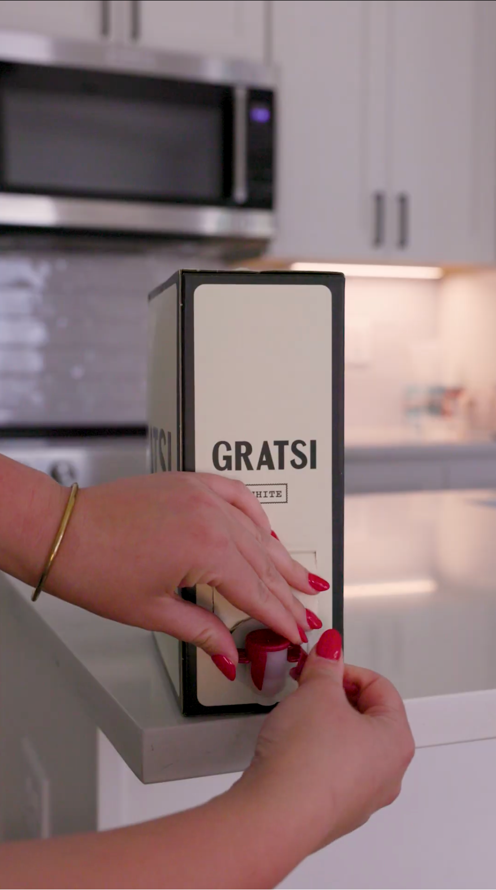 A person with red nail polish dispenses white wine from a Gratsi box on a kitchen counter, with white cabinets and a microwave in the background.