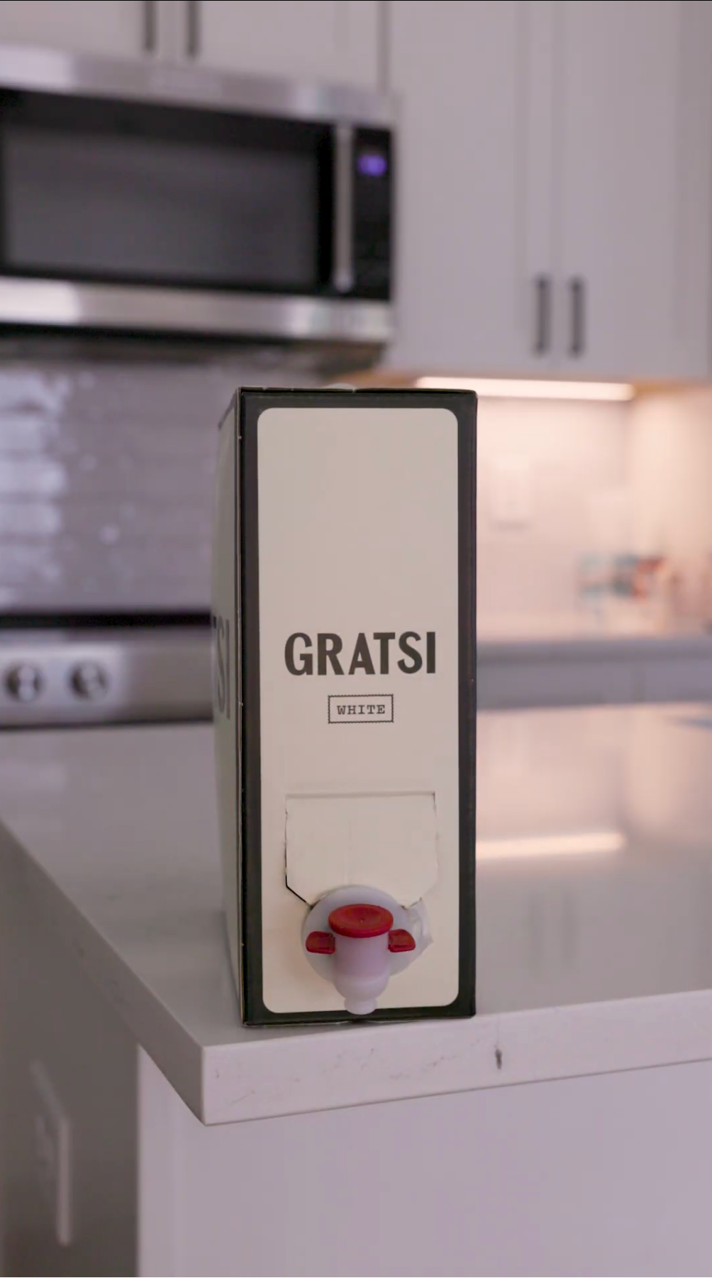 A boxed wine container labeled GRATSI WHITE sits on a white kitchen counter, with modern white cabinets and a microwave visible in the background.