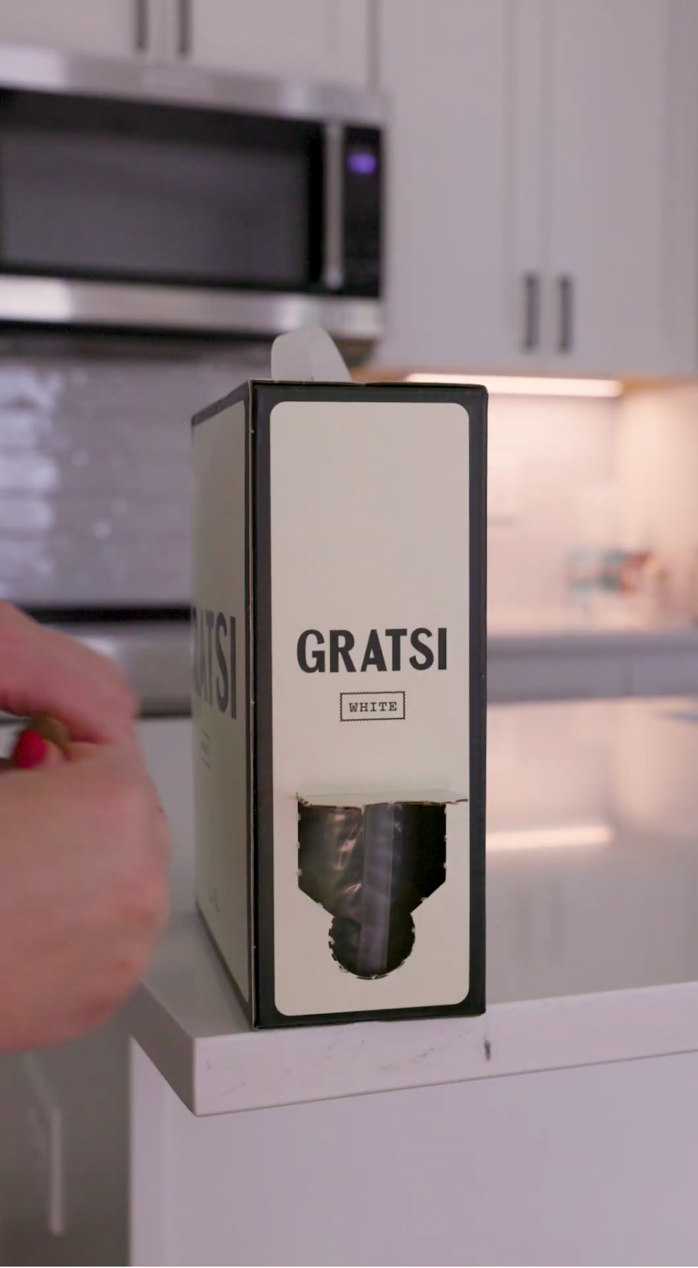 A hand reaches toward a box labeled GRATSI WHITE on a kitchen counter, with a spout visible at the bottom of the box. Kitchen appliances and cabinets are in the background.