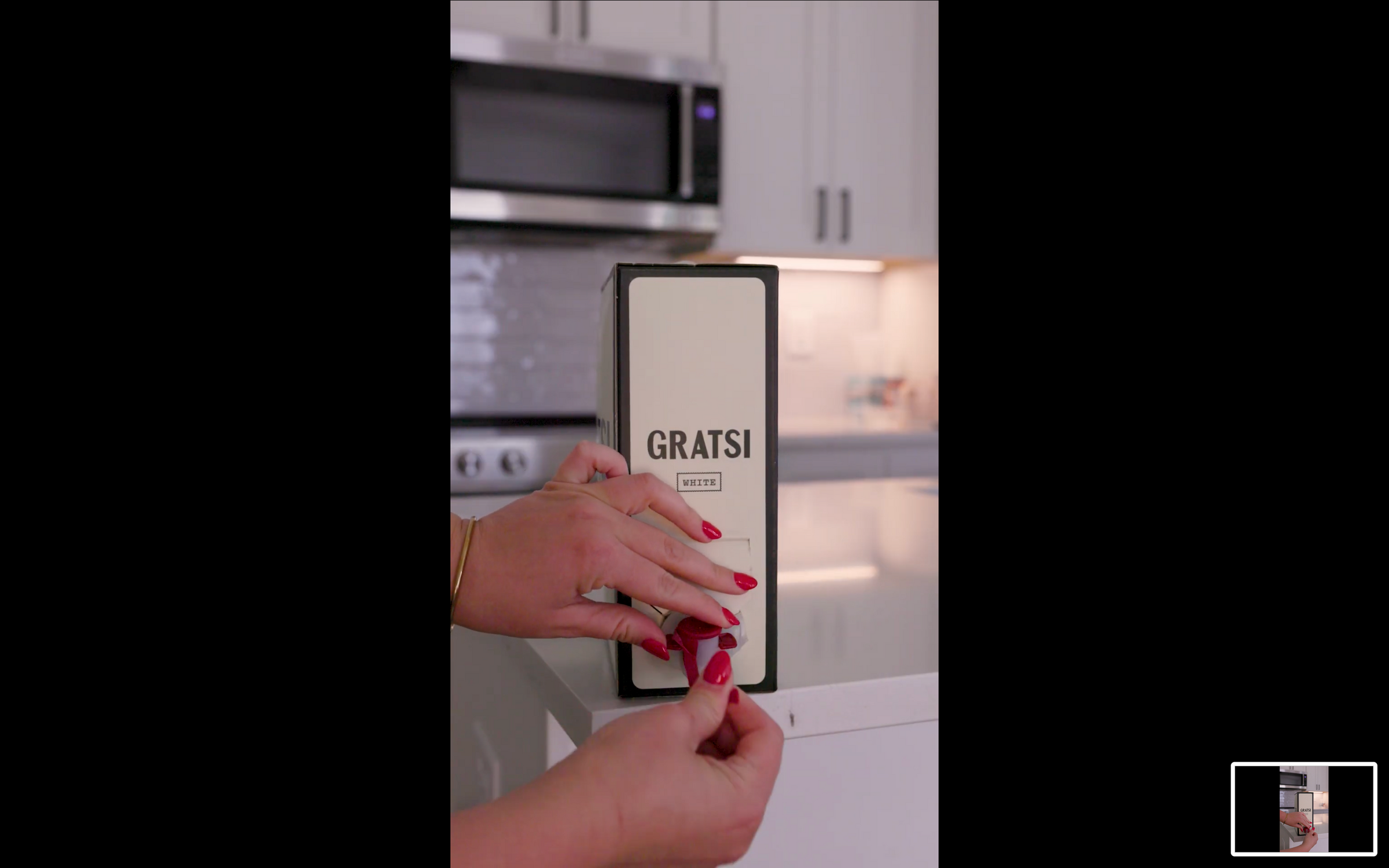 A person with red nail polish pulls a tab on a GRATS! branded box in a modern kitchen, with a stainless steel microwave and white cabinets in the background.