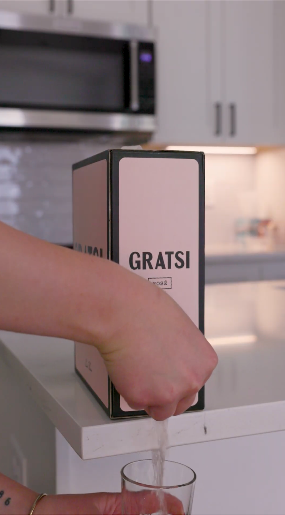 A person fills a glass from a boxed beverage labeled GRATSI on a white kitchen counter, with white cabinets and a microwave in the background.