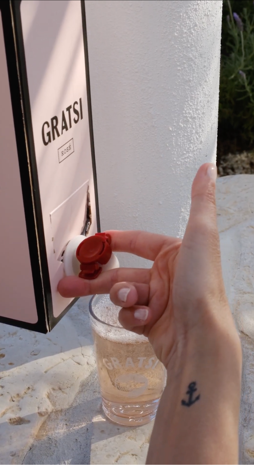 A hand with an anchor tattoo dispenses rosé wine from a pink Gratsi box into a branded glass, against a white textured background.