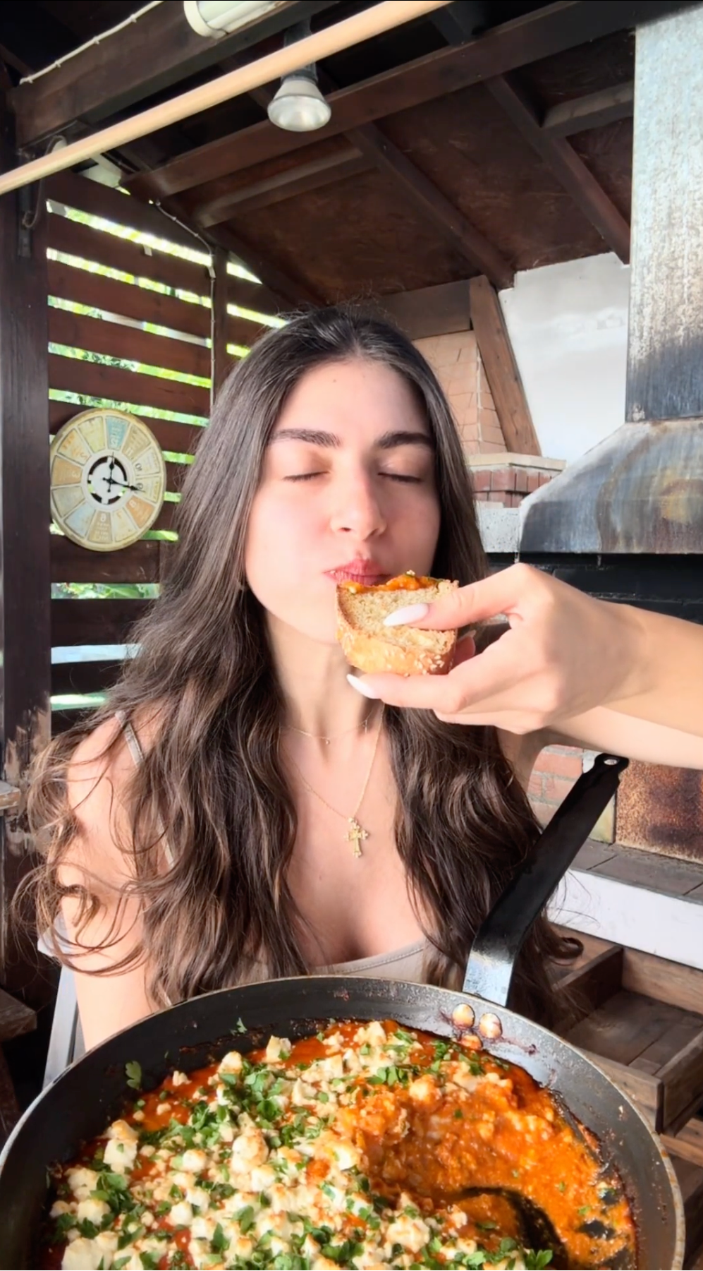 A woman with long brown hair sits at a table outdoors with her eyes closed, enjoying a bite of food from a piece of bread held by another hand. A skillet of food with herbs and cheese is in the foreground.
