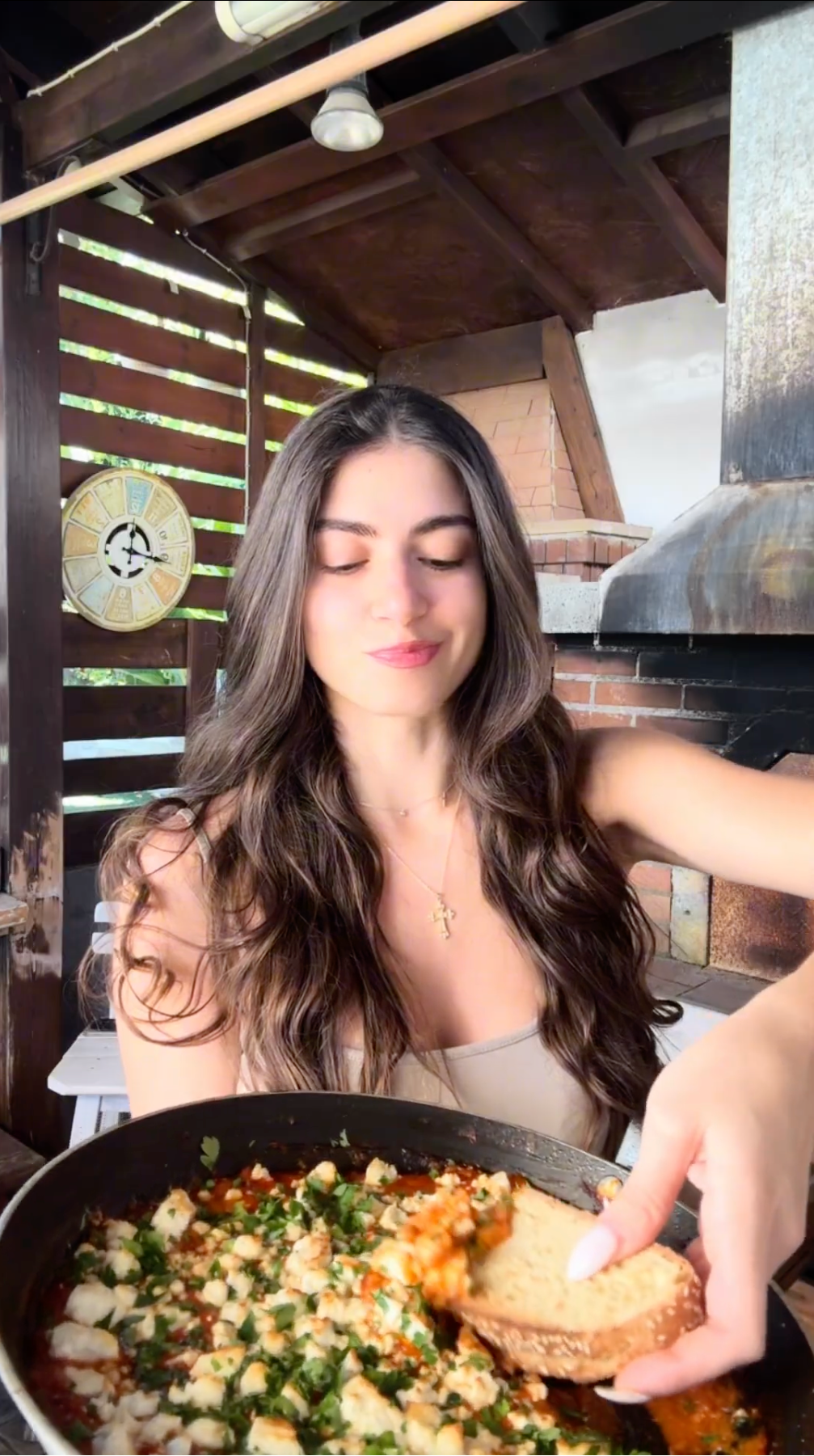 A woman with long brown hair is sitting outdoors by a brick oven, holding bread and dipping it into a pan of food topped with herbs and cheese. She appears to be enjoying the meal in a rustic setting.