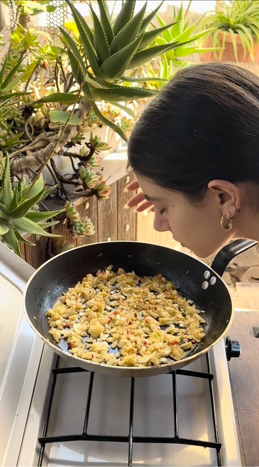 A woman leans over a pan of cooked food on a stove, smelling the aroma. The pan contains a mixture of vegetables and grains. There are several green potted plants in the background.