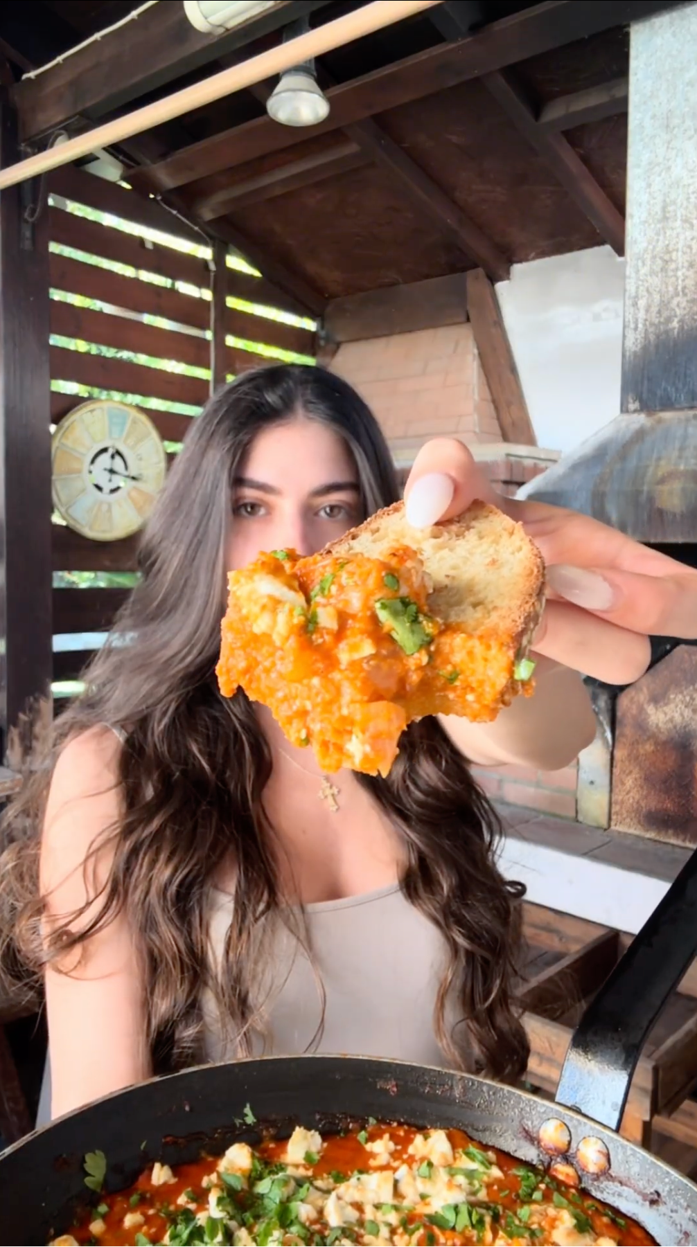 A woman with long brown hair holds up a piece of bread topped with a chunky, orange-red spread, in front of a pan of the same dish, outdoors near a wooden structure.