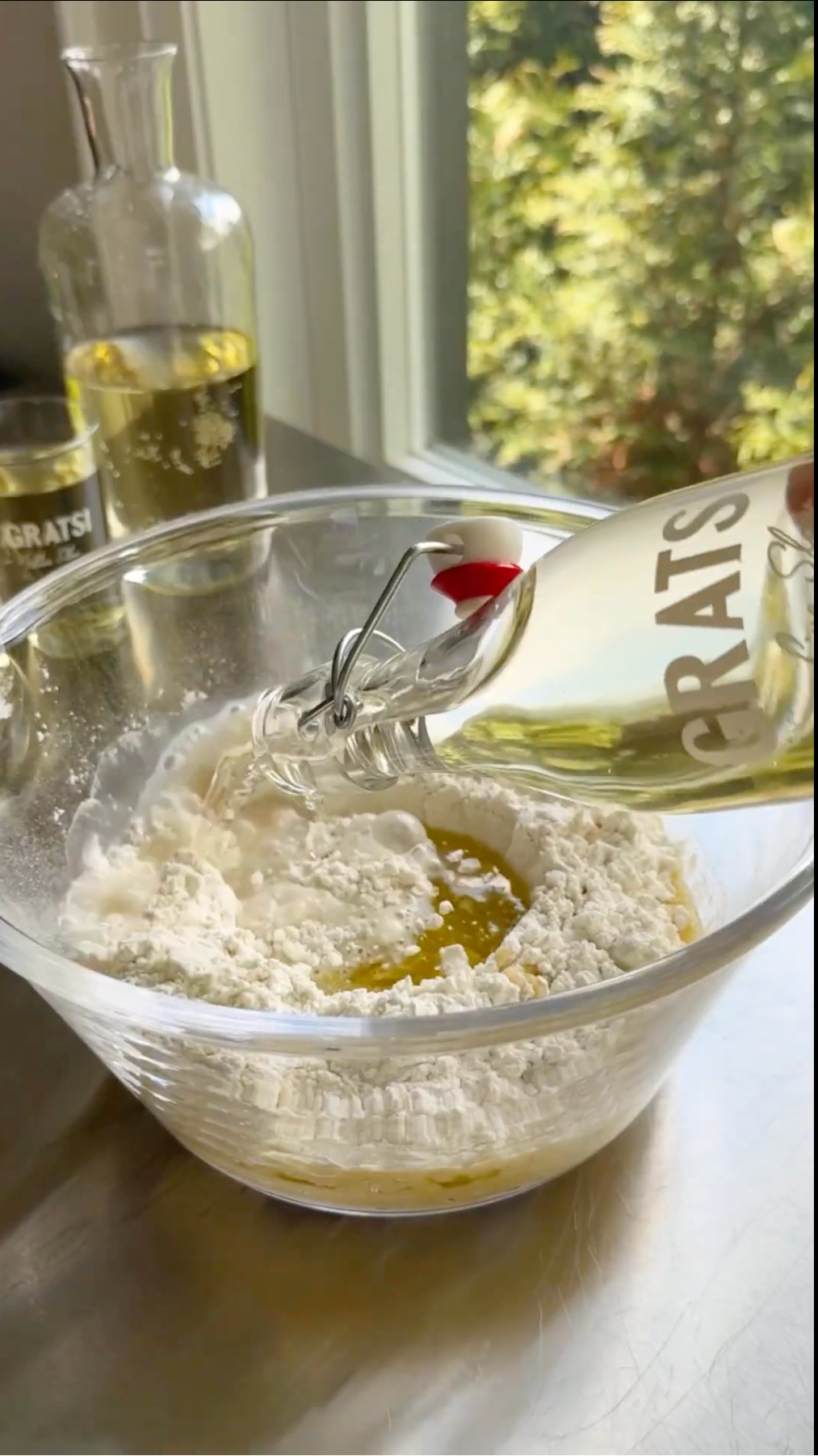 A hand pours clear liquid from a bottle labeled GRATS into a mixing bowl filled with flour and oil. A bottle and glass sit on the counter, and a window with greenery is in the background.