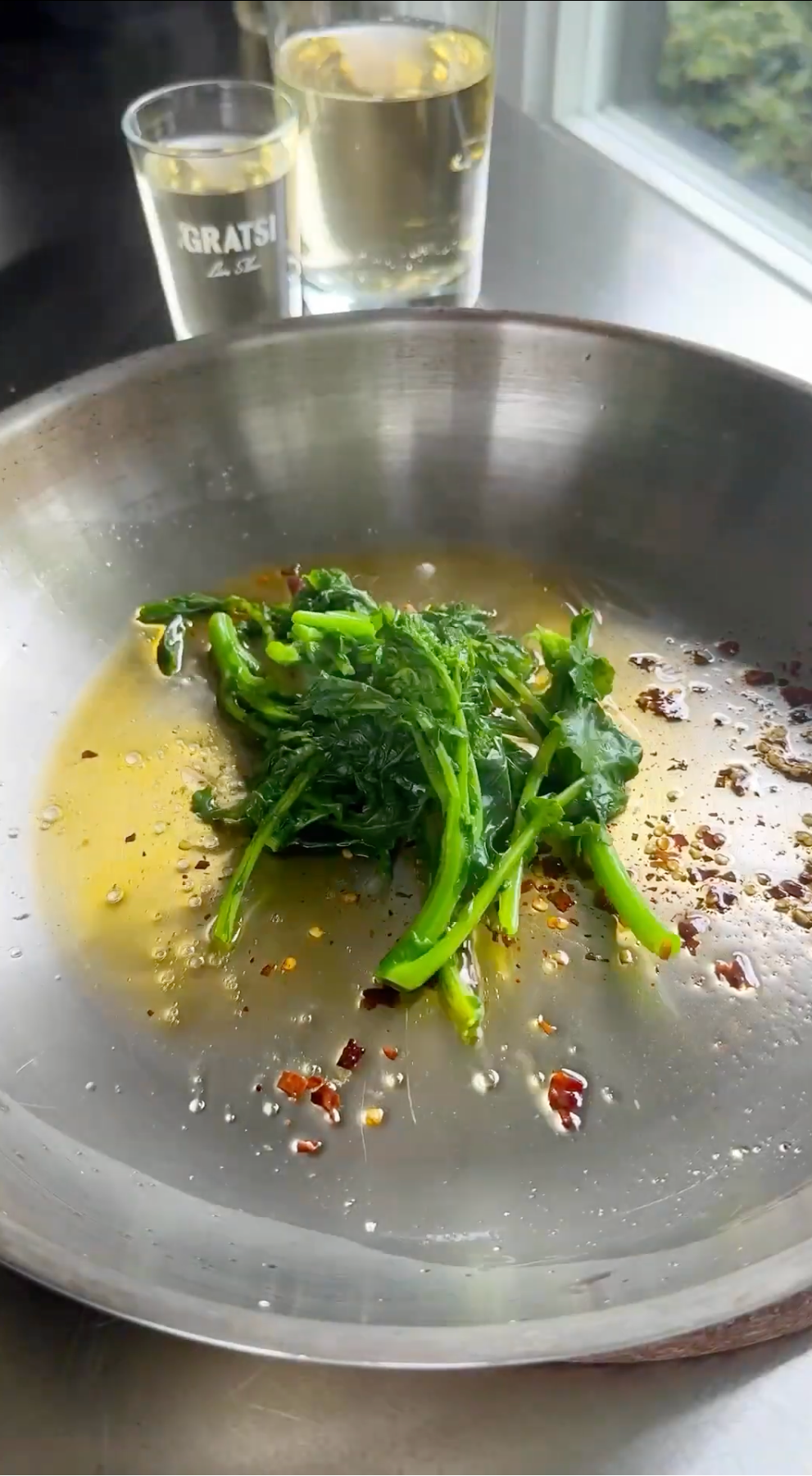 A close-up of sautéed greens with chili flakes and oil in a metal pan, with two glasses of white wine in the background near a window.