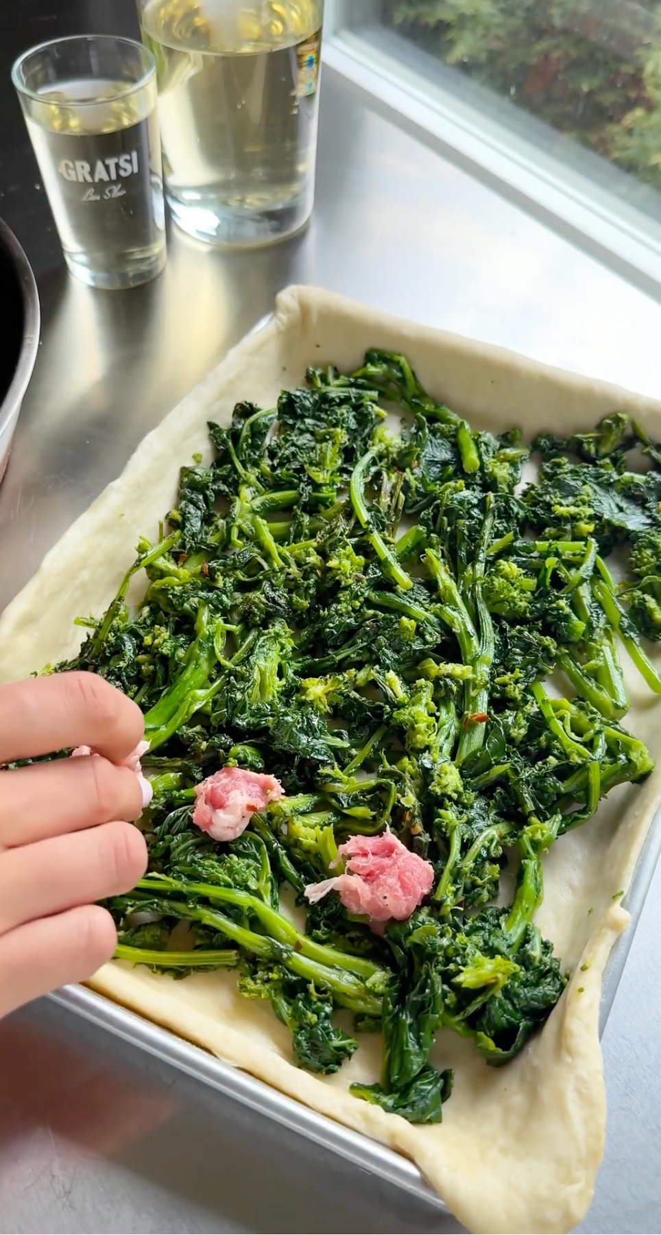 A hand adds small pieces of sausage to a rectangular sheet of dough topped with sautéed greens, likely broccoli rabe, on a metal baking tray. A glass of white wine sits nearby on the counter.