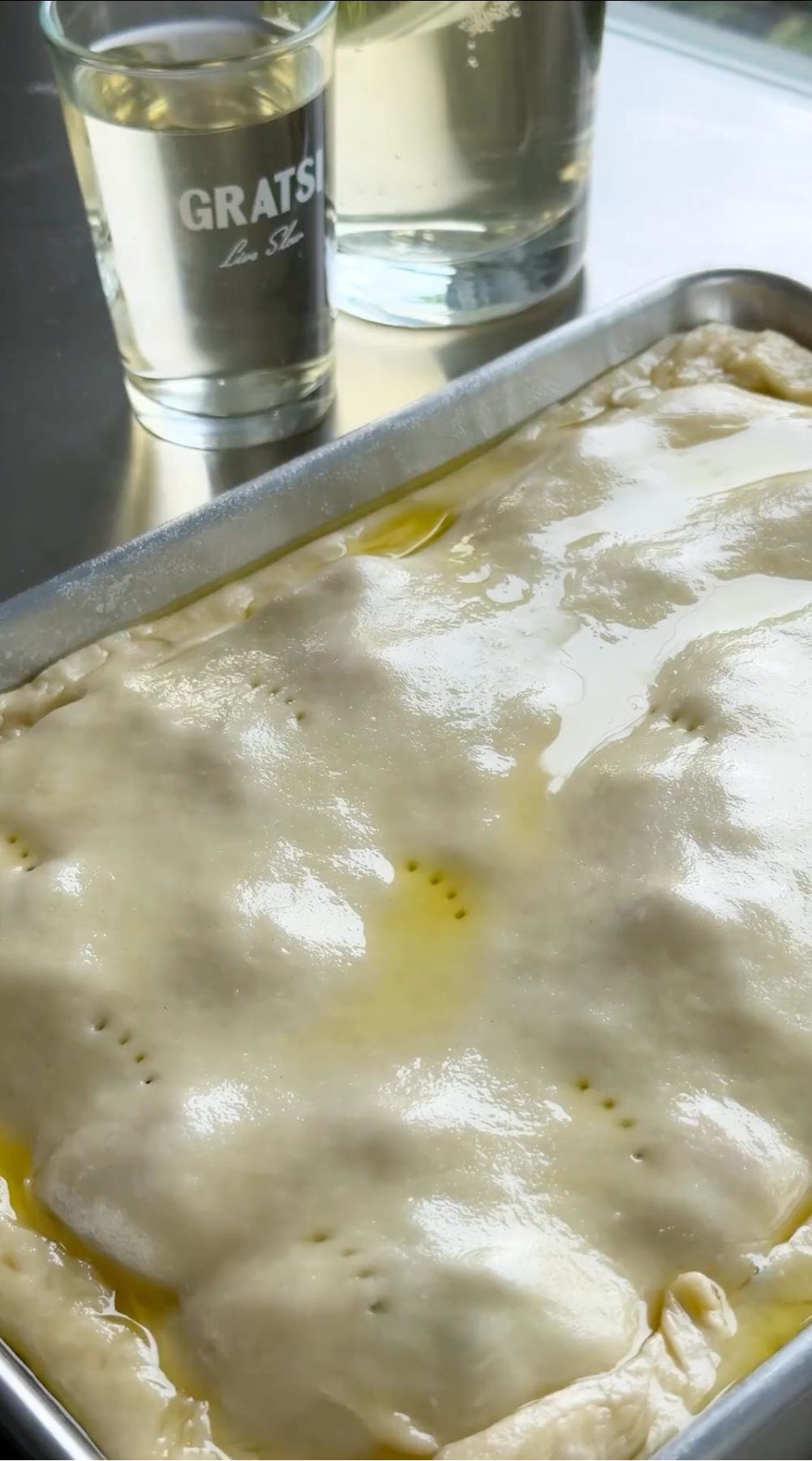 A rectangular pie with a golden, glossy crust sits on a metal tray next to two glasses filled with a clear, light-colored beverage. The pie crust is slightly puffy with small fork holes.