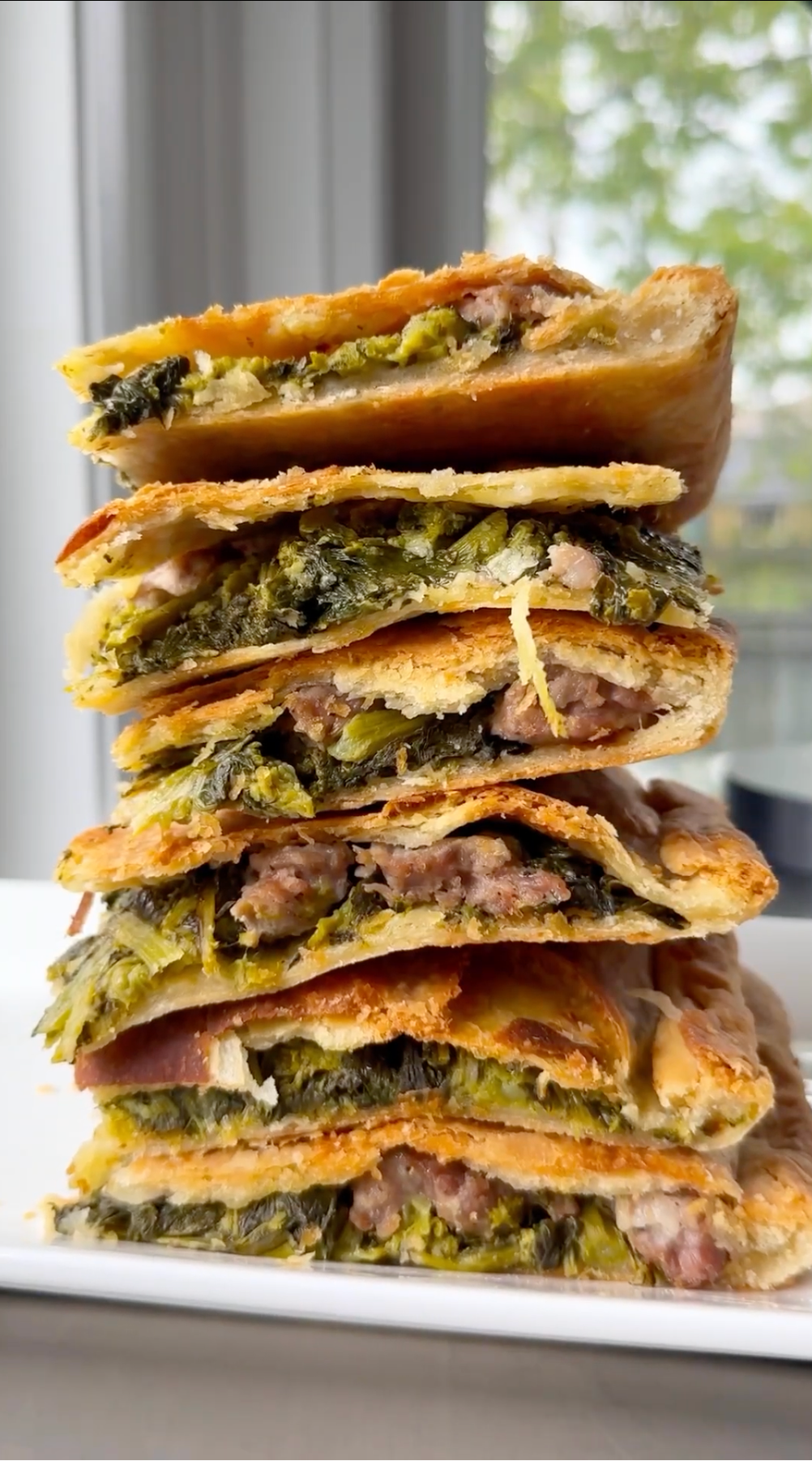 A stack of golden-brown, flaky pastry slices filled with sausage and leafy greens is neatly arranged on a white plate, with a blurred window and greenery in the background.