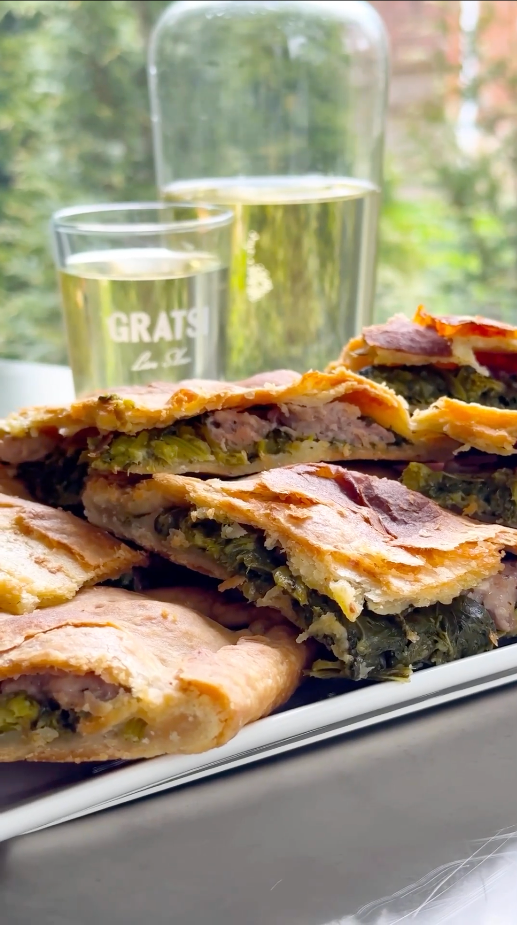 A plate of golden-brown, flaky pastries filled with greens and possibly meat, stacked in layers. In the background, there are two glasses of water, one with the word GRATS printed on it, and a blurred green outdoor scene.