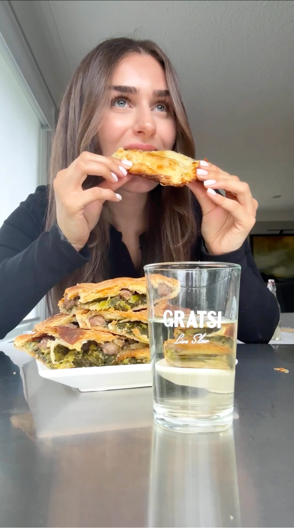 A woman with long brown hair and a black top smiles while eating a slice of pizza. In front of her are stacked pieces of pizza and a clear glass of water labeled “GRATSI”.