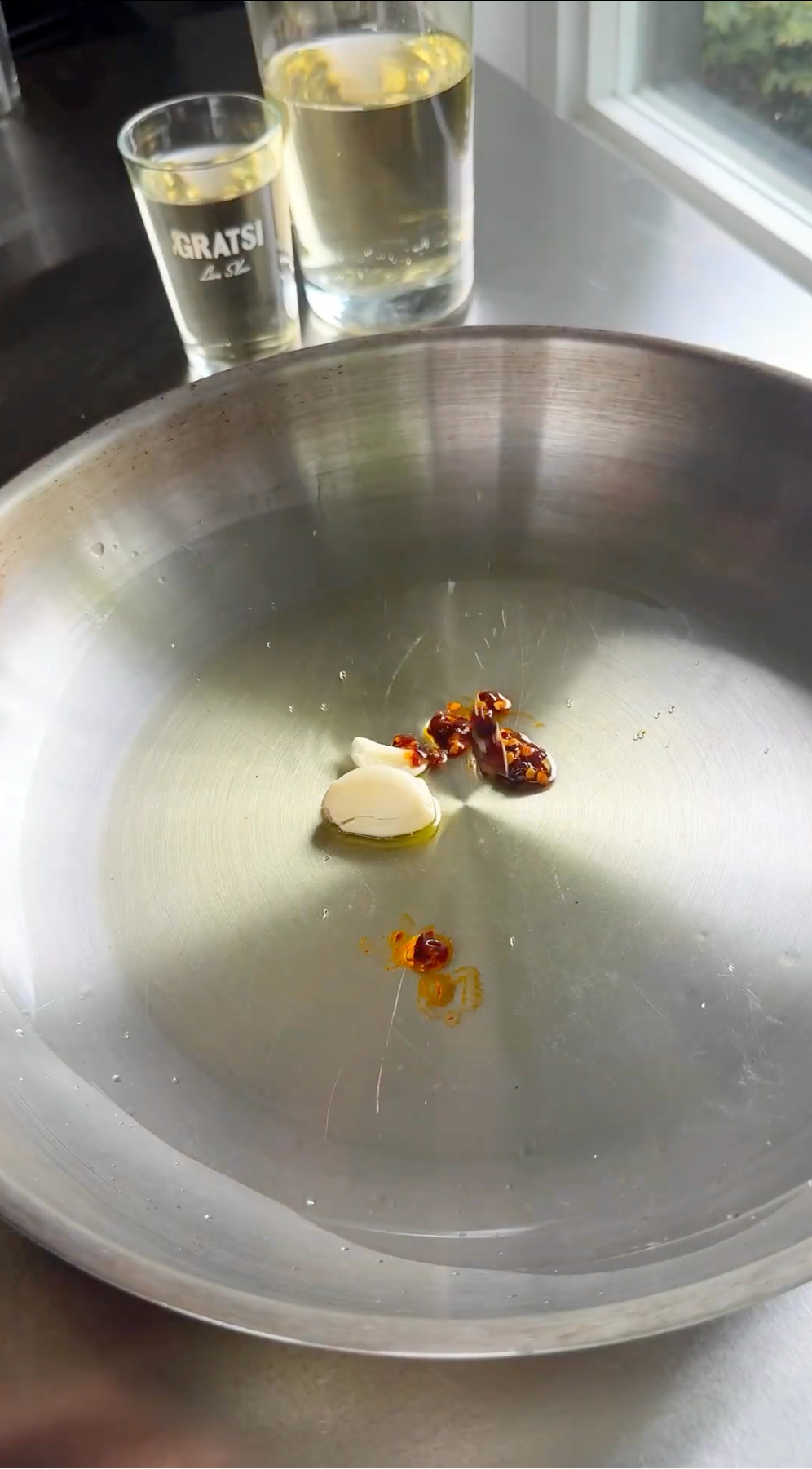 A stainless steel pan with a clove of garlic, a small amount of chili paste or sauce, and oil inside. In the background, there are two glasses—one half-filled with white wine and one with a clear liquid.
