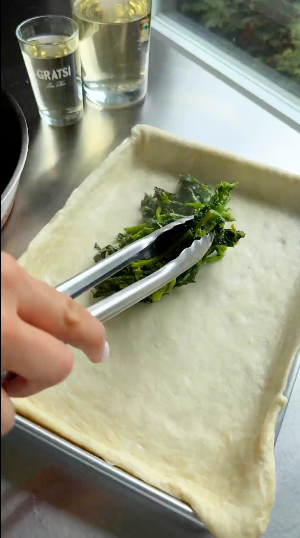 A hand uses tongs to place sautéed greens onto a rectangular sheet of dough in a baking pan. A glass of white wine and a shot glass are on the counter nearby.