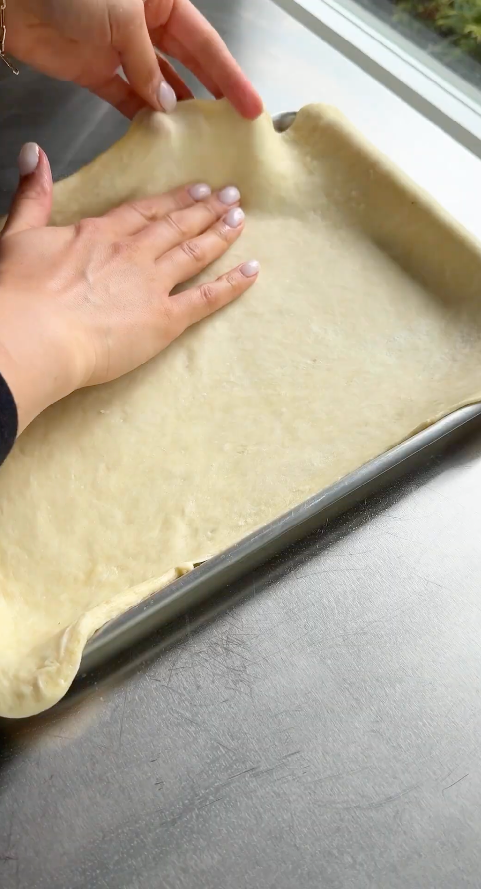 Hands pressing pizza dough evenly into a rectangular baking pan on a metal countertop, smoothing it out to fit the edges.