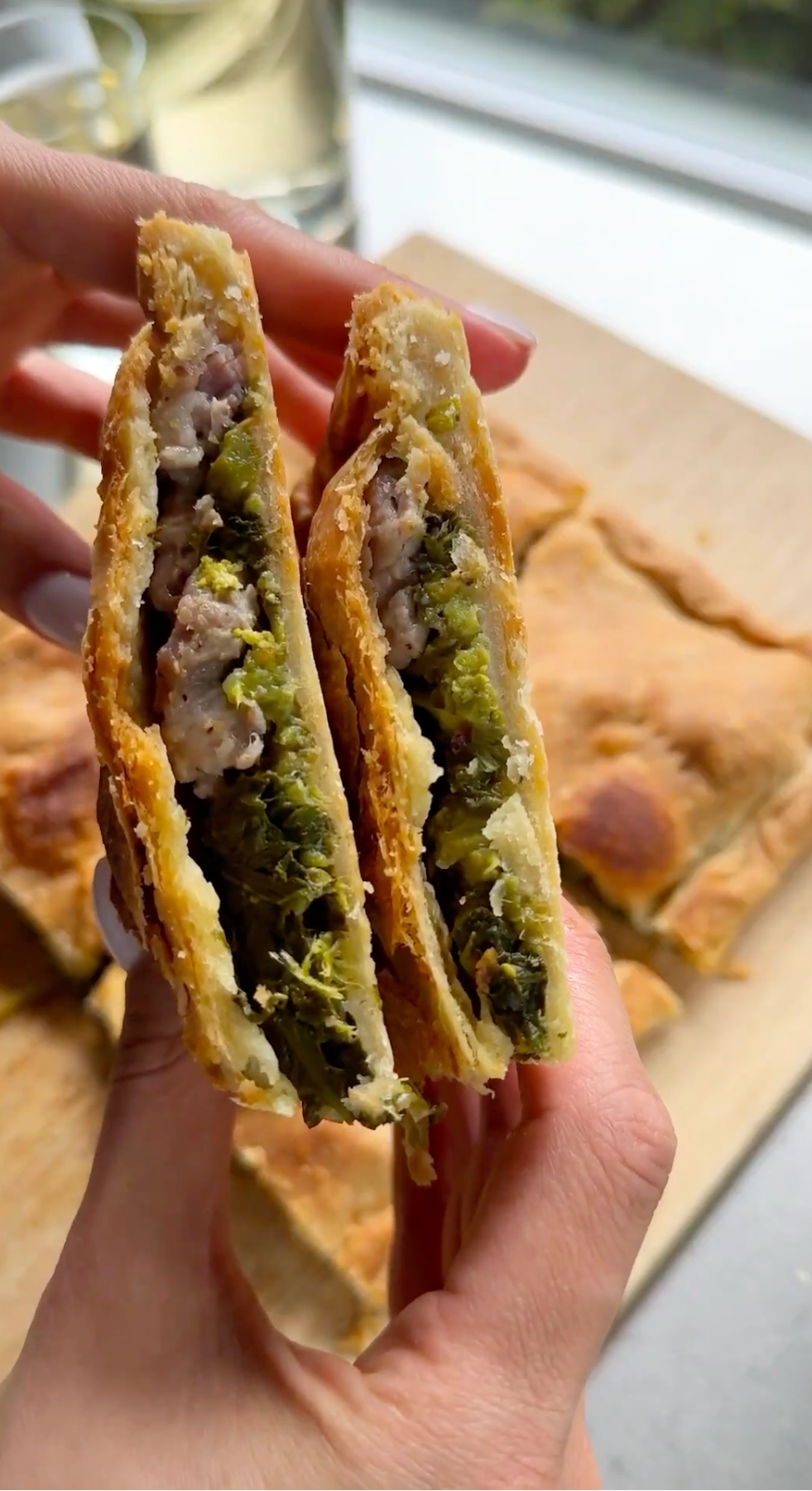 Two hands hold two halves of a savory pastry filled with minced meat and green vegetables, showing the flaky crust and the filling. More square-cut pieces of the same pastry are on a wooden board in the background.