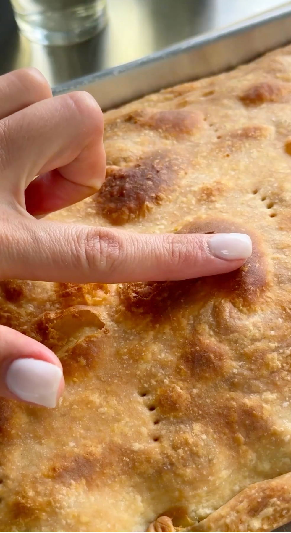 A hand with light pink polished nails gently presses a finger into the golden-brown, bubbly surface of a freshly baked pie.