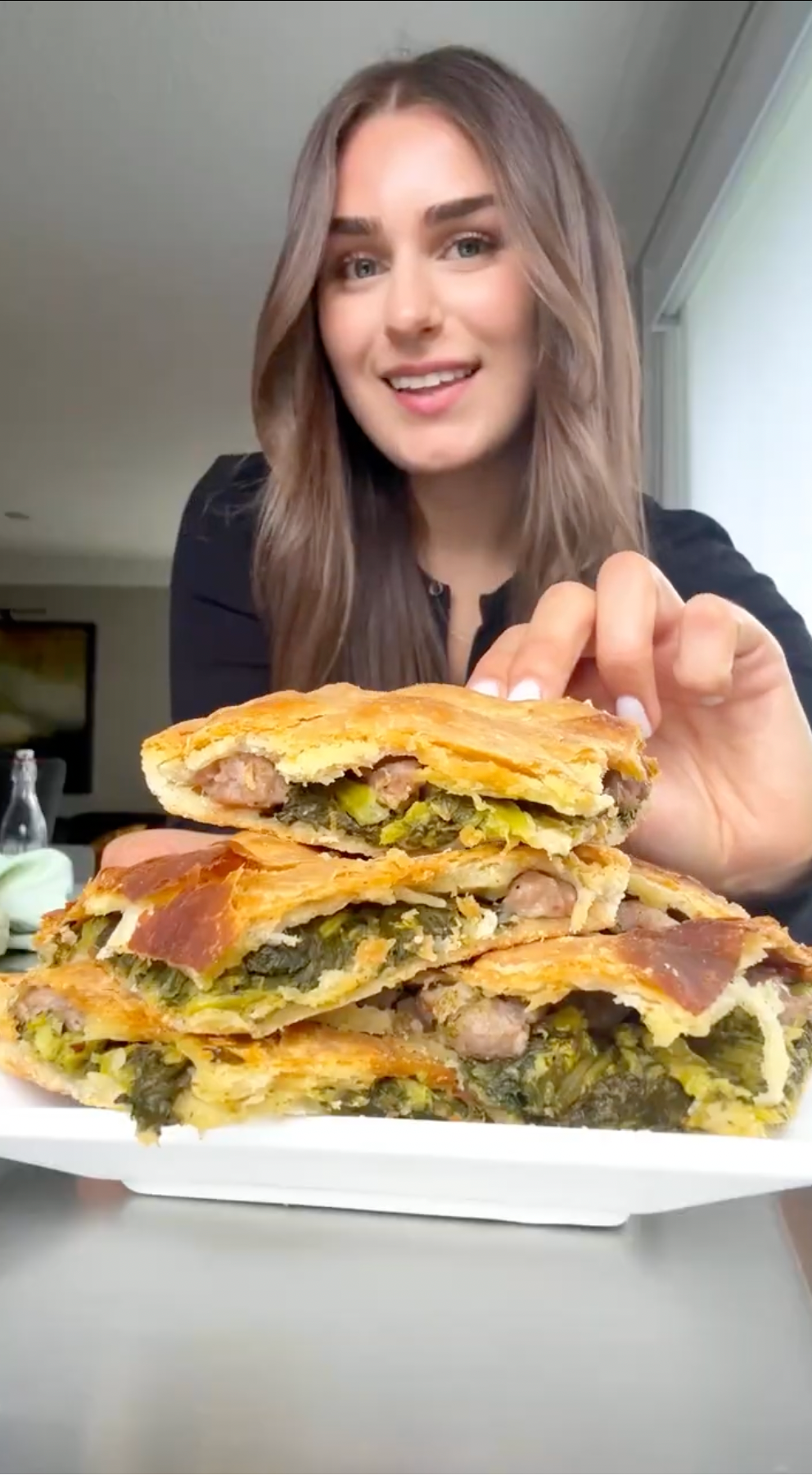 A smiling woman with long brown hair holds a plate stacked with slices of savory pie filled with greens and sausage, in a well-lit indoor setting.