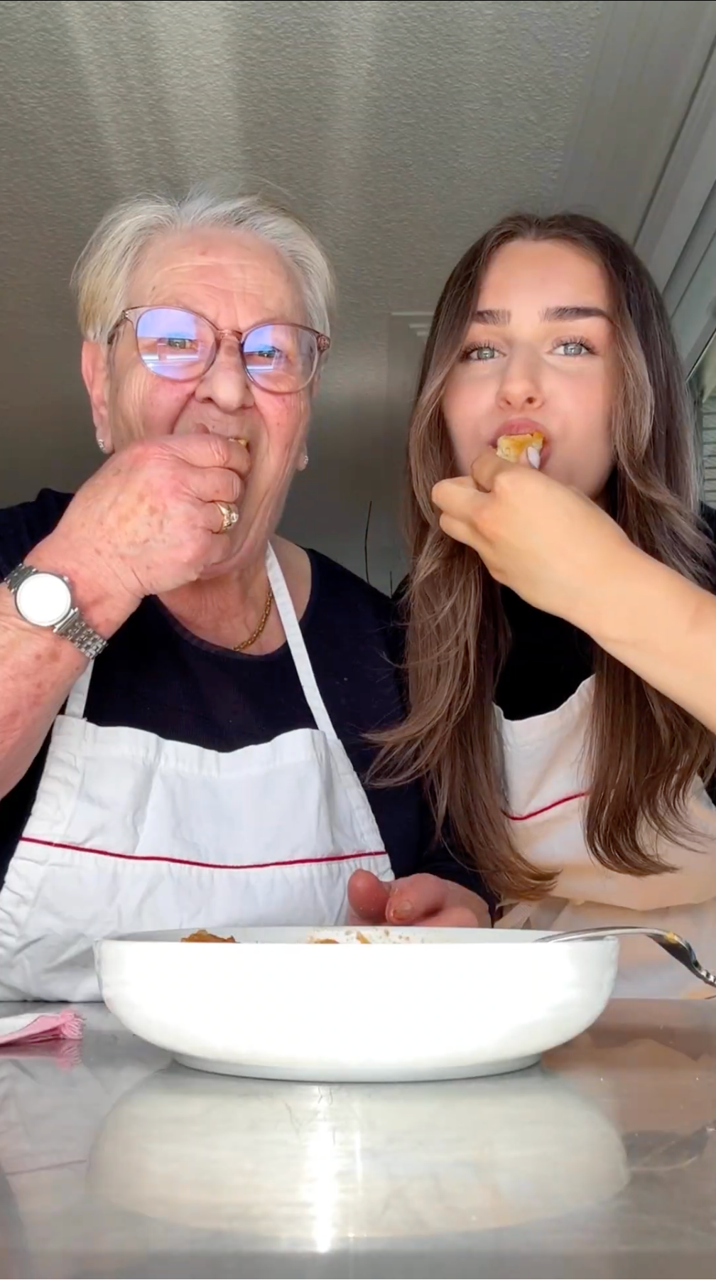 An older woman and a young woman, both wearing aprons, sit side by side and smile at the camera while eating food together from a white dish. They appear to be enjoying a meal in a bright kitchen.