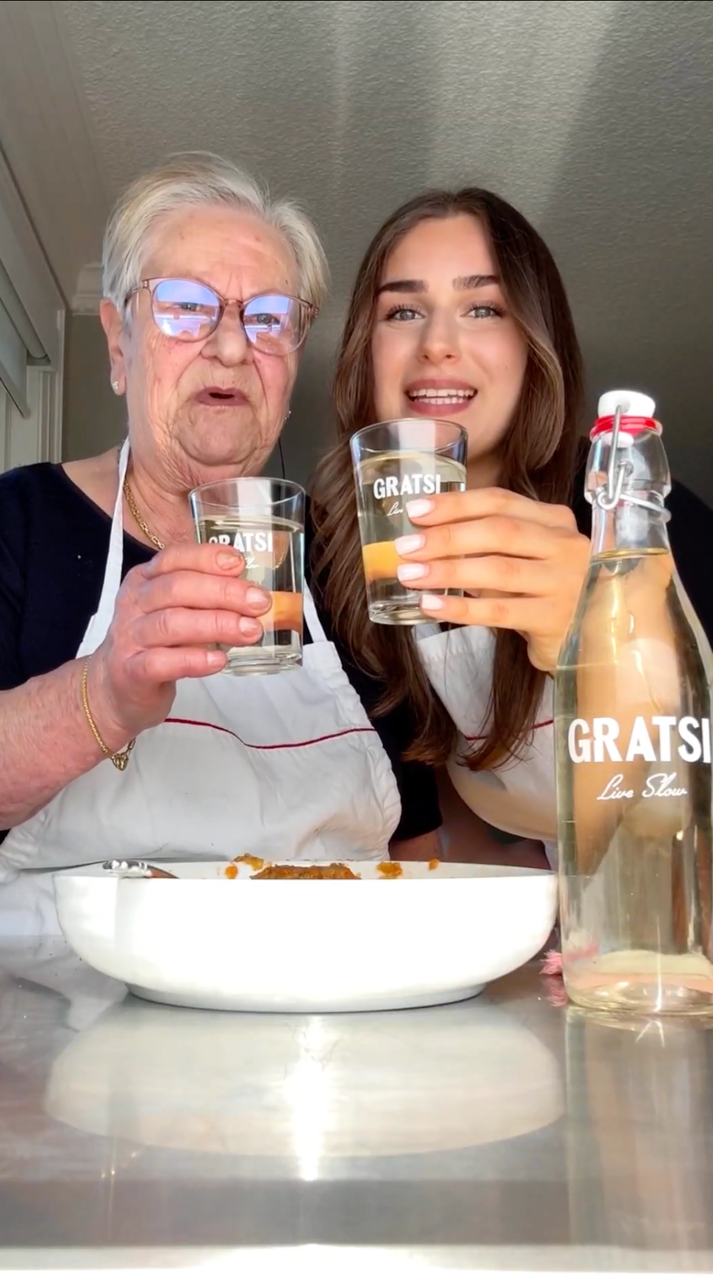 An elderly woman and a young woman smile and toast with glasses labeled GRATSI in a bright kitchen. A bottle and a plate of food are on the table in front of them.