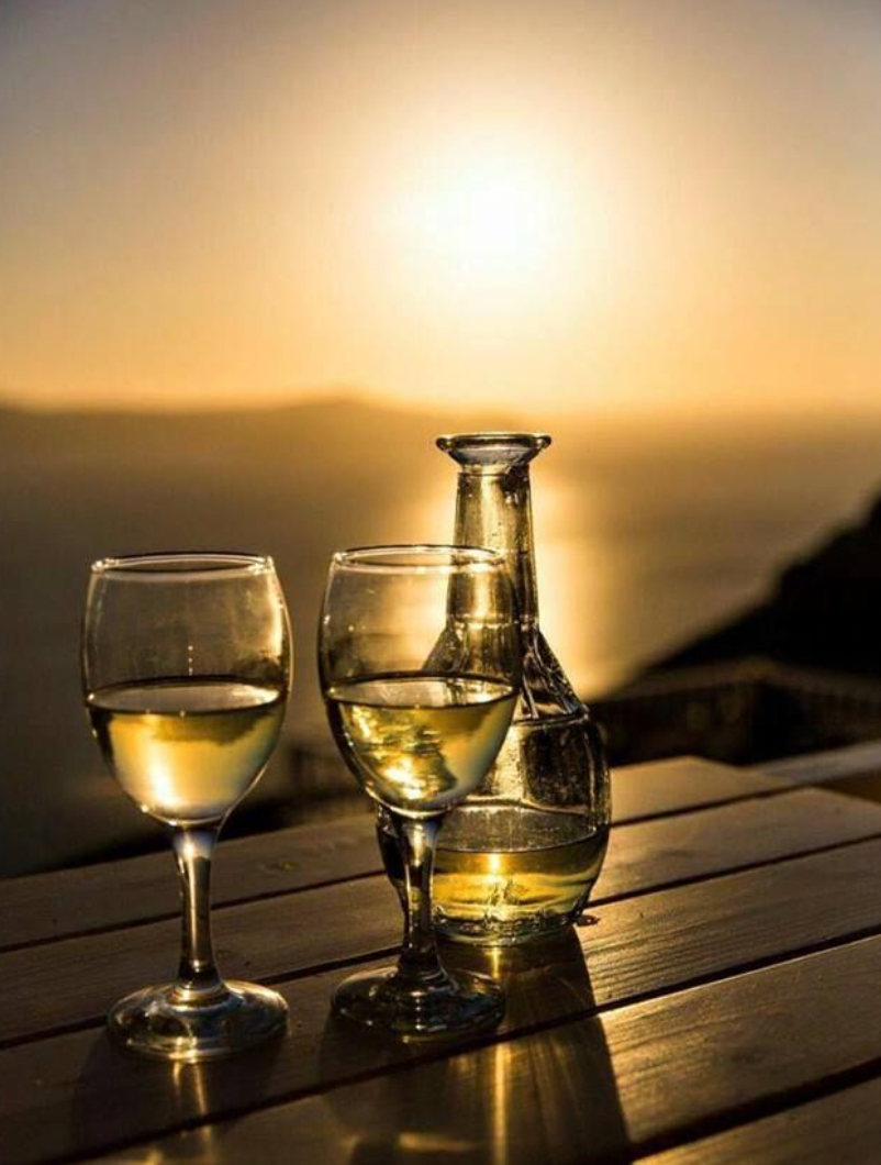 Two glasses and a carafe of white wine sit on a wooden table, overlooking a calm body of water at sunset with the golden sun reflected on the waters surface.