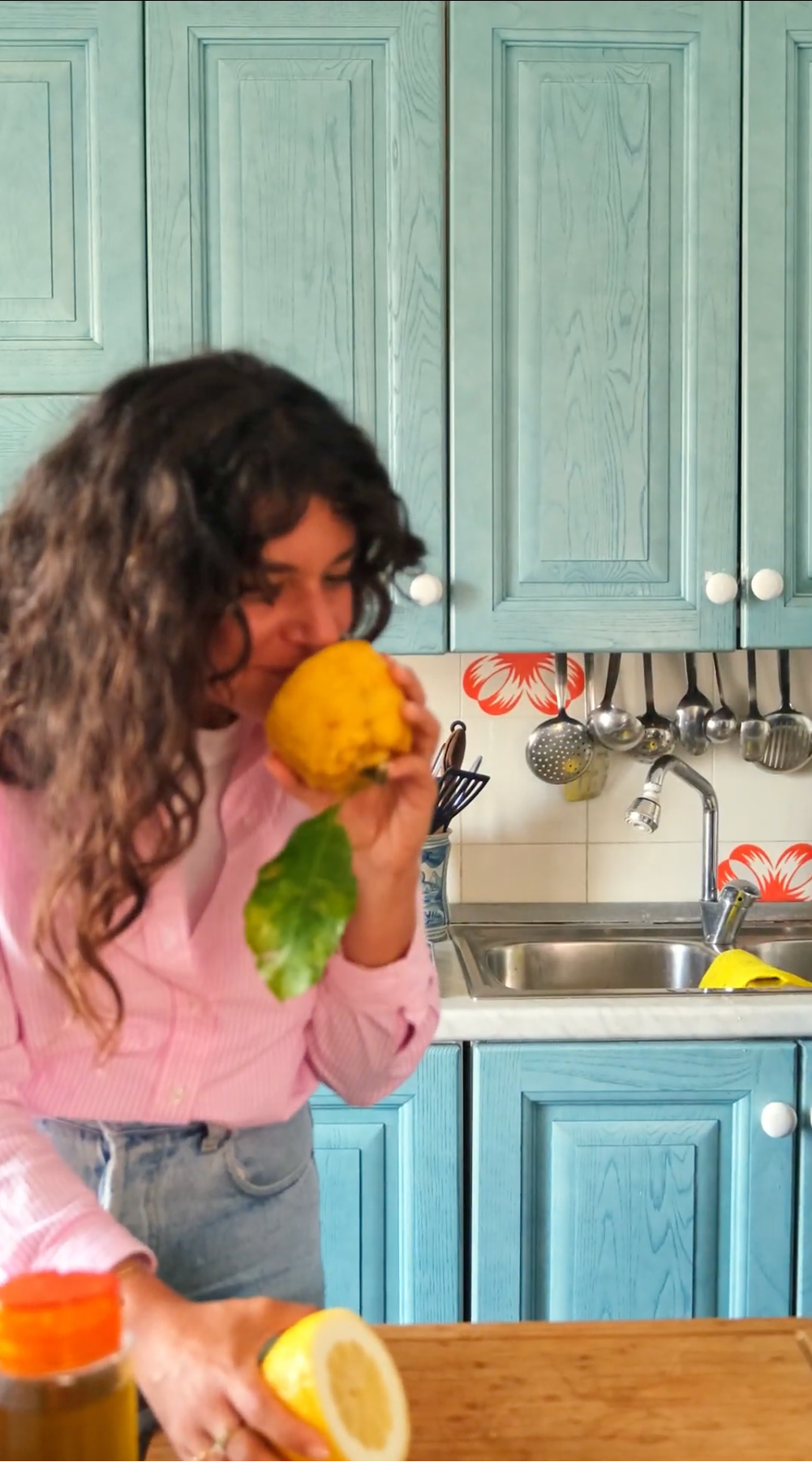 A woman with long curly hair, wearing a pink shirt and jeans, holds and smells a large yellow fruit with a green leaf in a kitchen with blue cabinets and utensils hanging by the sink.