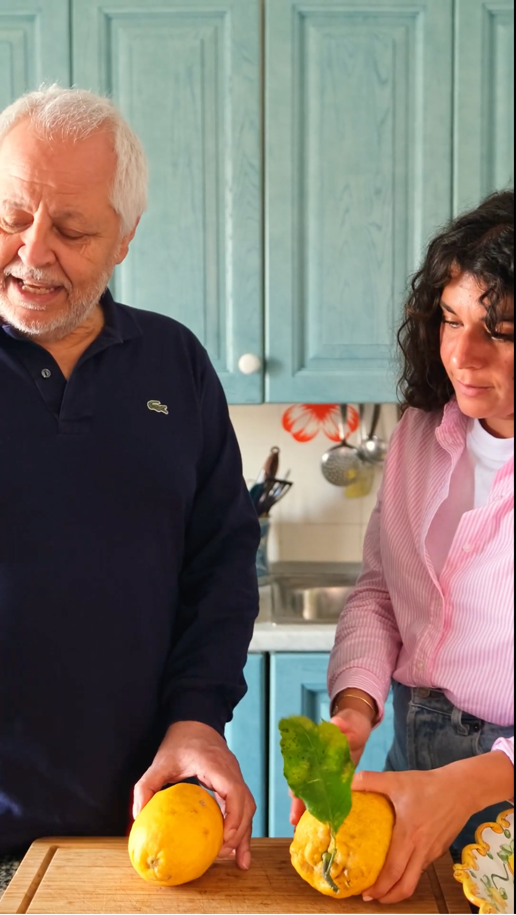 An older man and a younger woman stand in a kitchen holding large yellow fruits. The man smiles while the woman looks at the fruit. Blue cabinets and utensils are visible in the background.