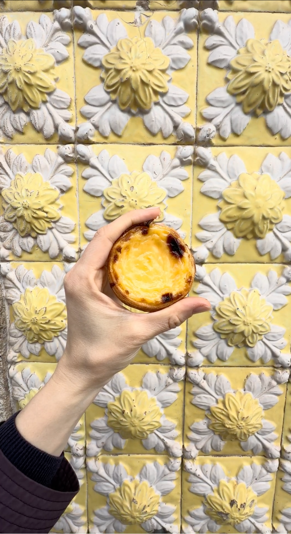 A hand holds up a pastel de nata (Portuguese custard tart) in front of a wall decorated with yellow and white floral ceramic tiles.